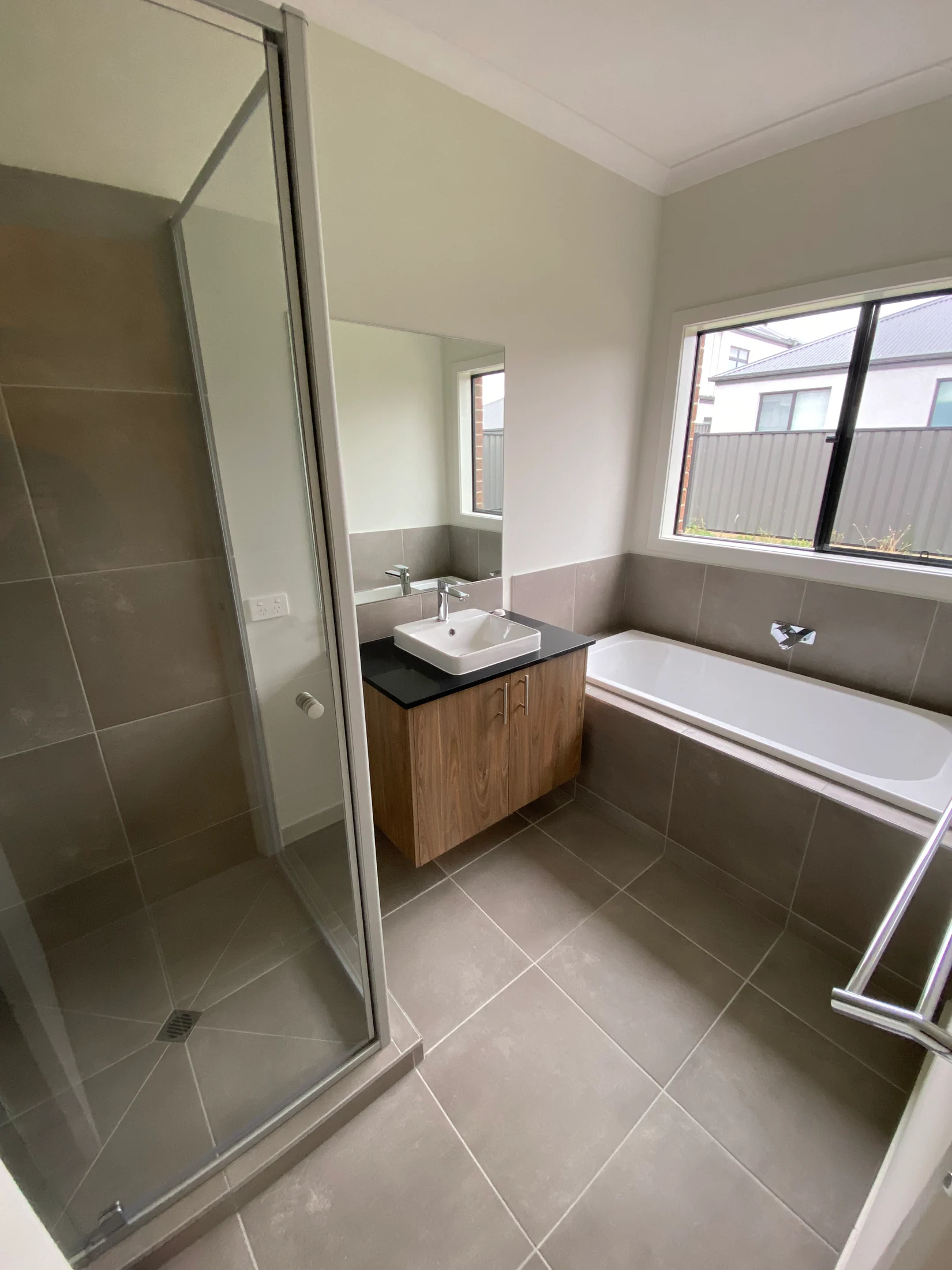 Modern bathroom with a glass shower, wood vanity with a sink, and a bathtub. Neutral tiles and a window create a calm atmosphere.