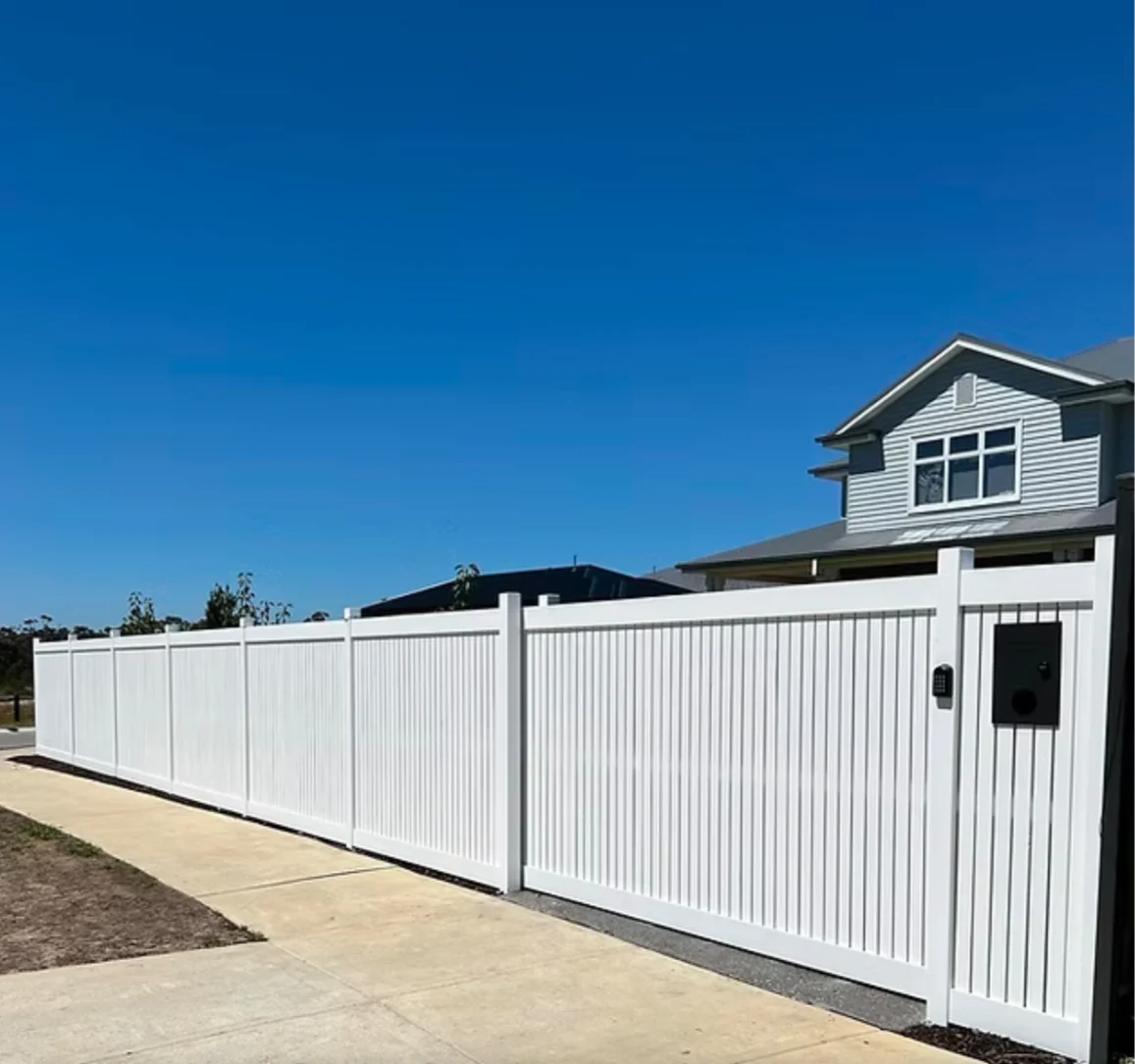 White fence and concrete path in front of a gray house under a clear blue sky. Simple and peaceful suburban setting.