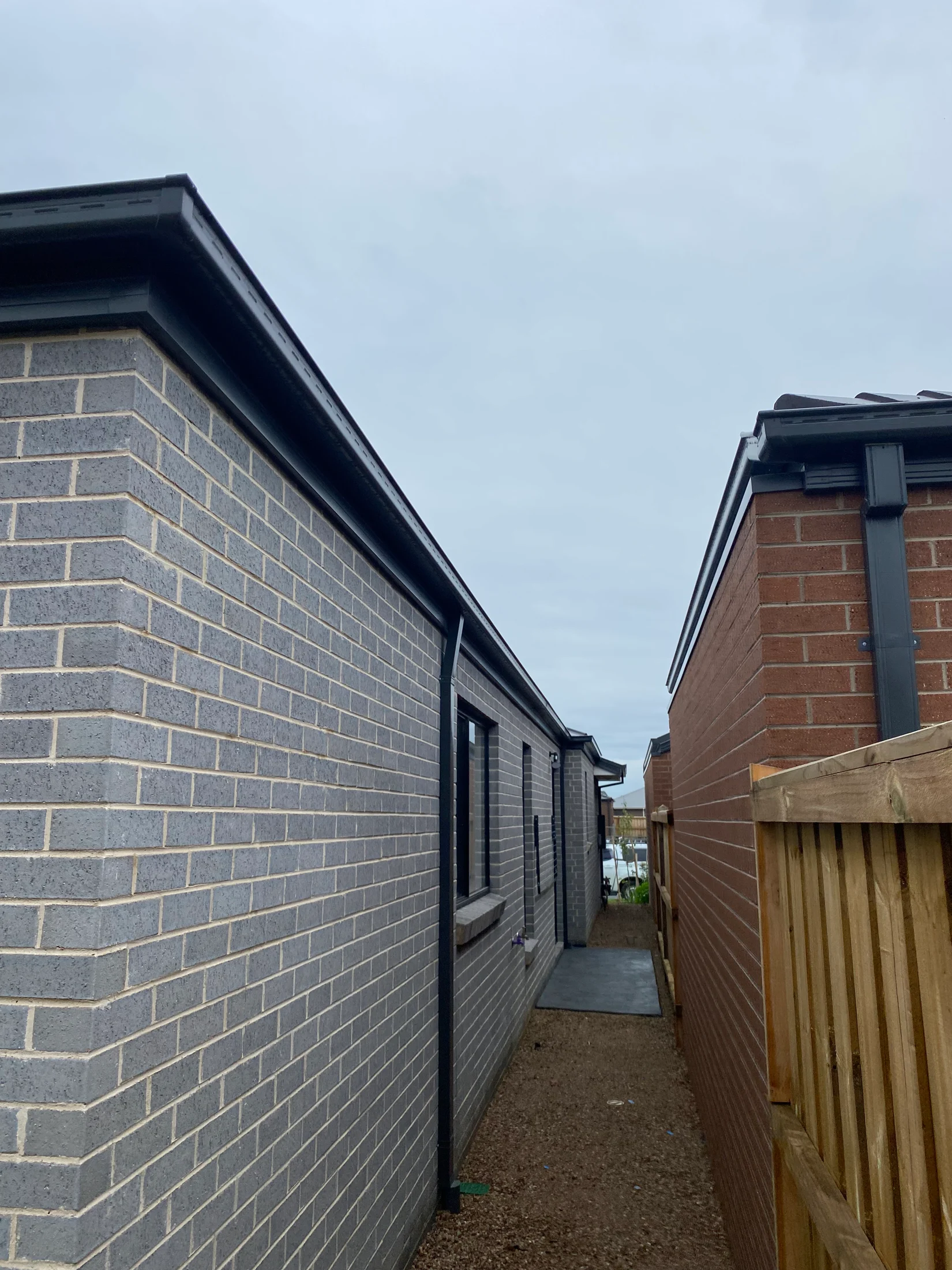Narrow alley between a grey brick building and a red brick house with wooden fencing. Overcast sky above, creating a calm atmosphere.