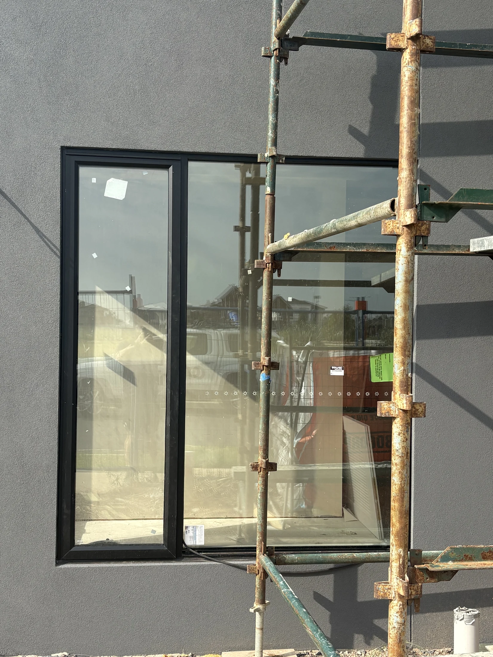 Window with black frame on a gray wall, adjacent to rusty scaffolding. Reflects construction site with a mix of sunny and shadowed areas.