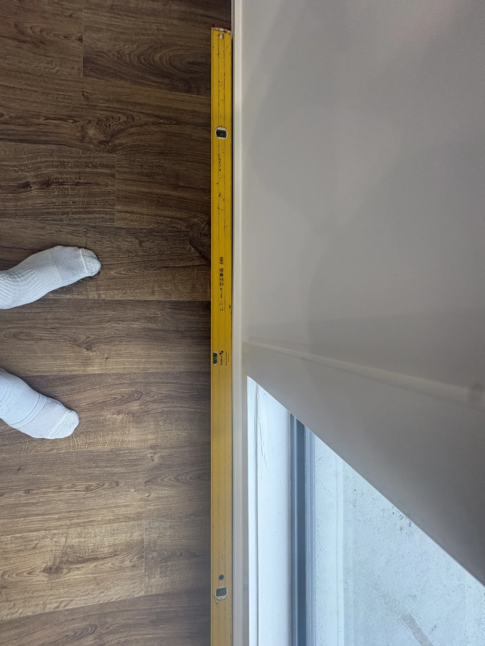 Yellow level on wood floor next to a white wall and window. A person in white socks stands nearby.
