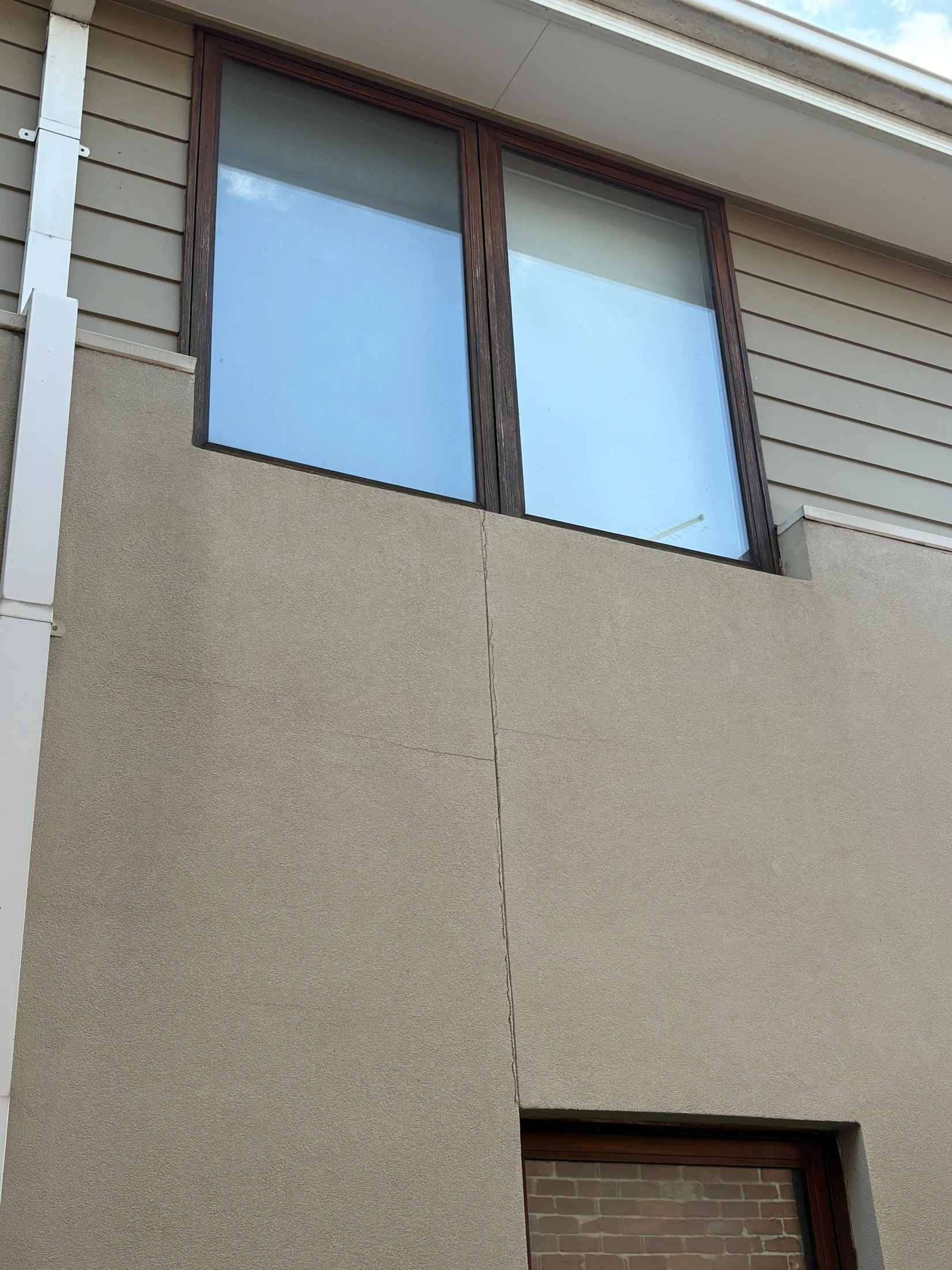 Facade of a beige house with a vertical crack, wooden-framed windows, and siding. The sky is partly visible, conveying a neutral mood.