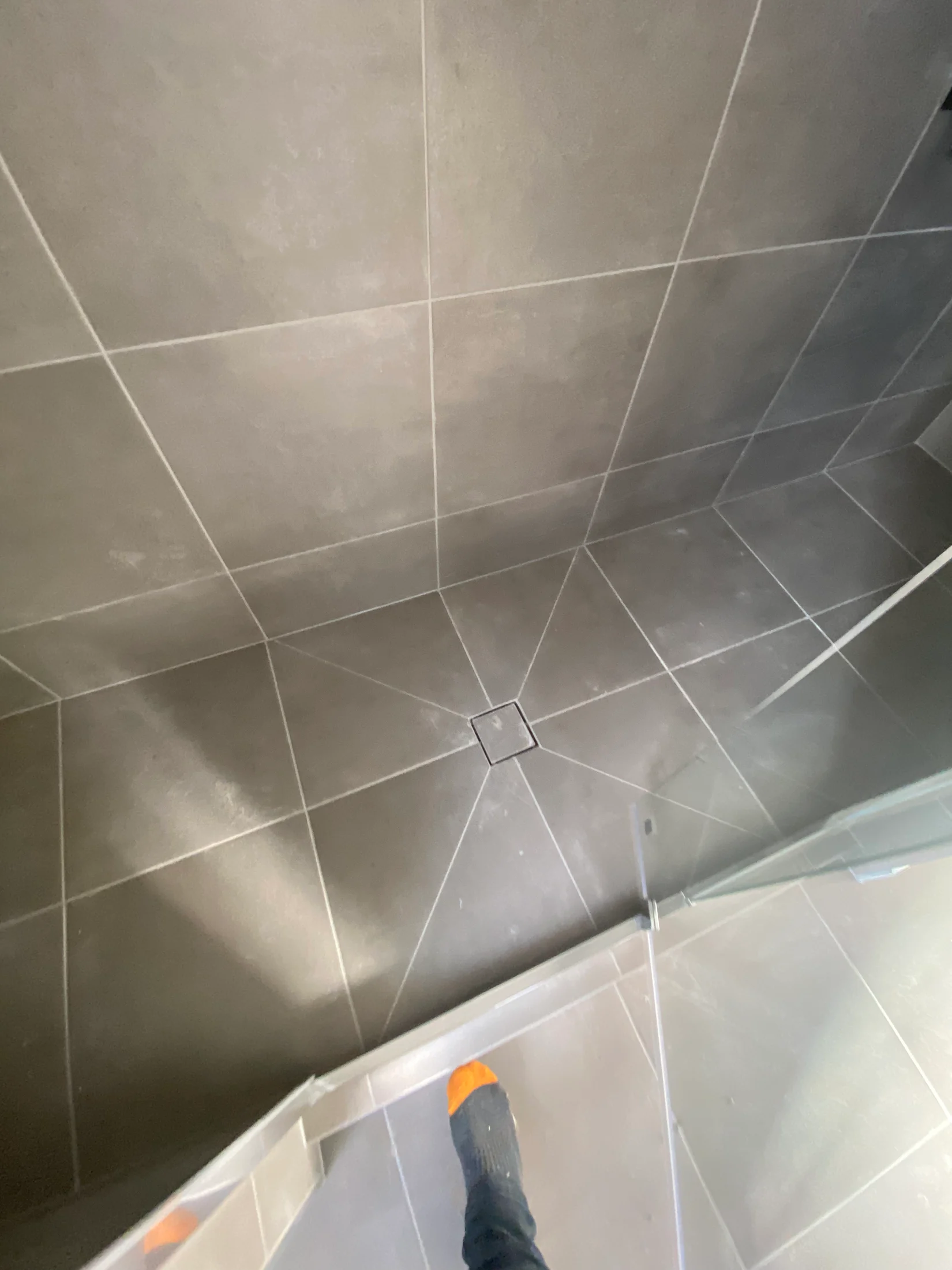 A foot in an orange-tipped sock steps onto a grey tiled shower floor. The tiles have a geometric pattern with subtle reflections.