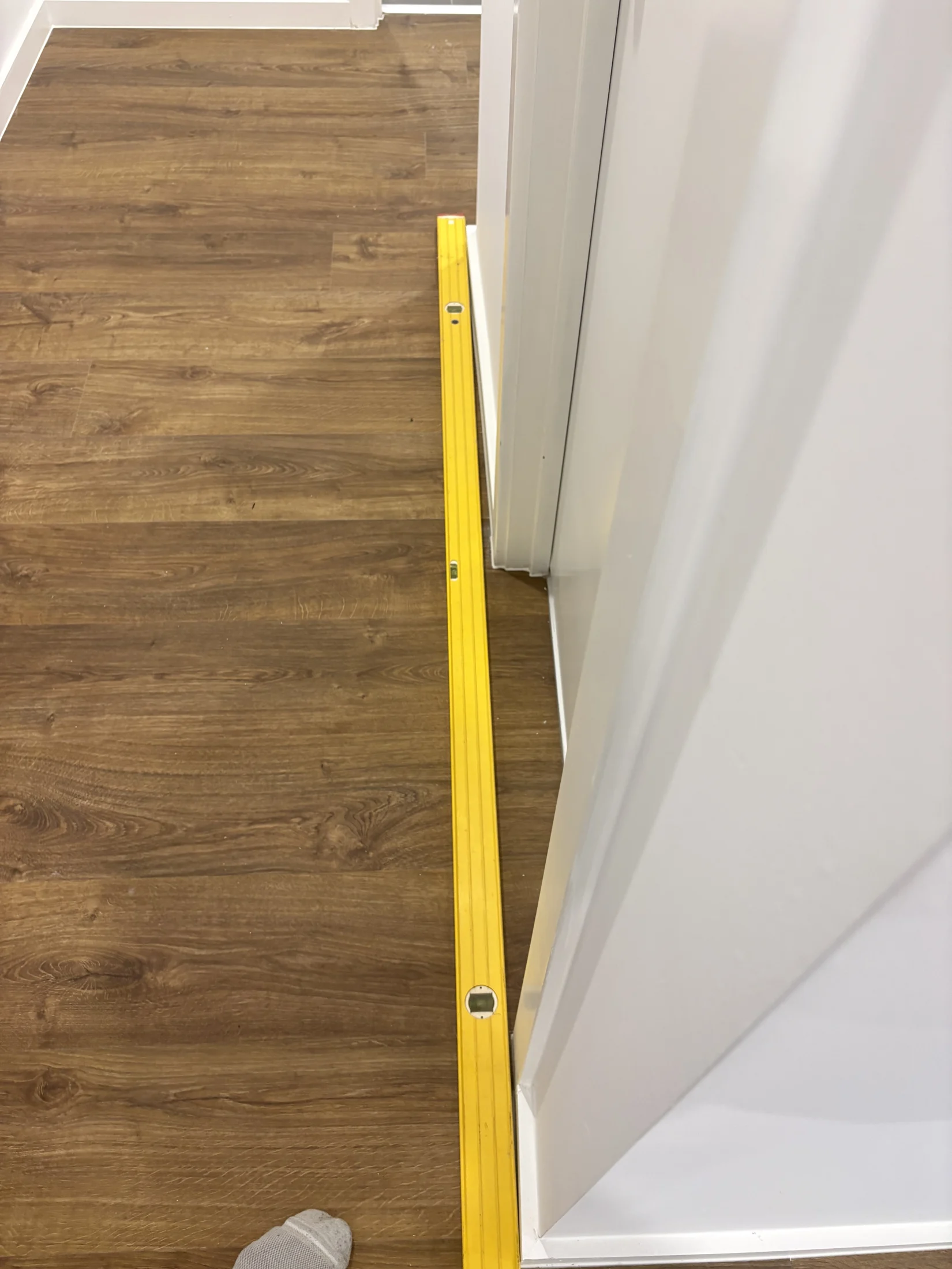 Yellow level on a wooden floor next to a white wall. Sock-covered foot visible. Bright lighting, no text.