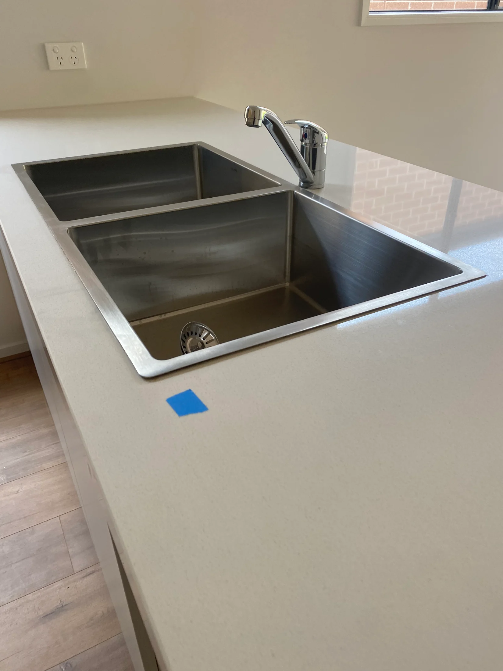 Stainless steel double sink on a beige countertop, chrome faucet, blue tape on surface, wood floor, and wall socket in background.