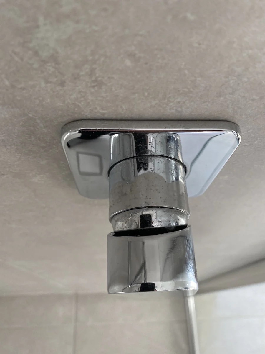 Close-up of chrome shower head in a bathroom with beige tile background. The shower head is unlit with visible reflections.