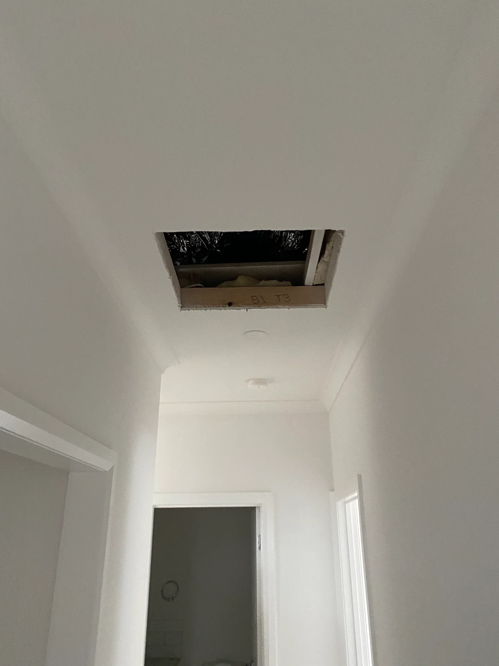 A hallway with a rectangular ceiling hole exposing insulation and ductwork. The walls and ceiling are white with no visible text.