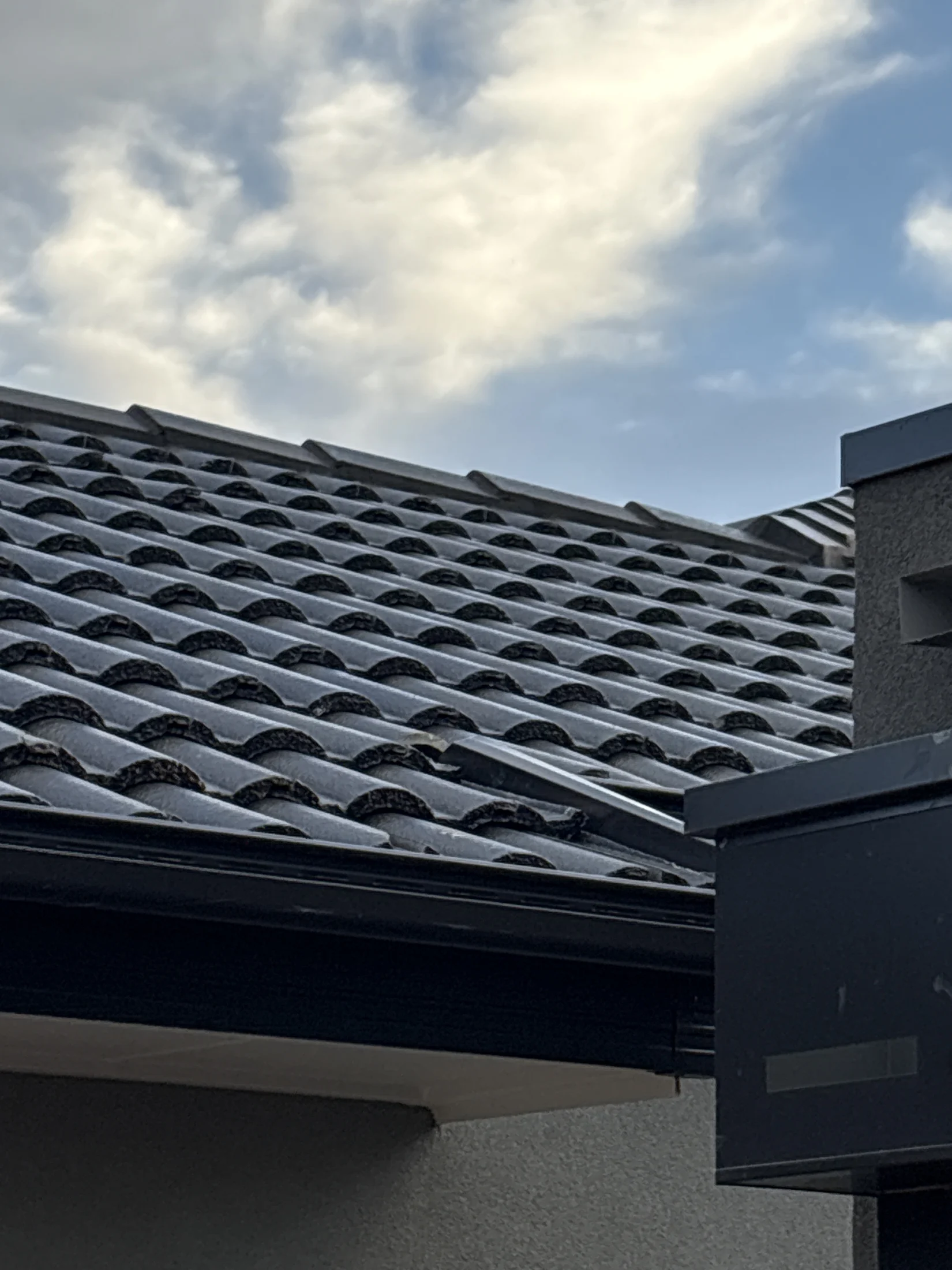A tiled roof with solar panels under a partly cloudy sky. The roof has a dark, shiny appearance, and the sky adds a serene atmosphere.