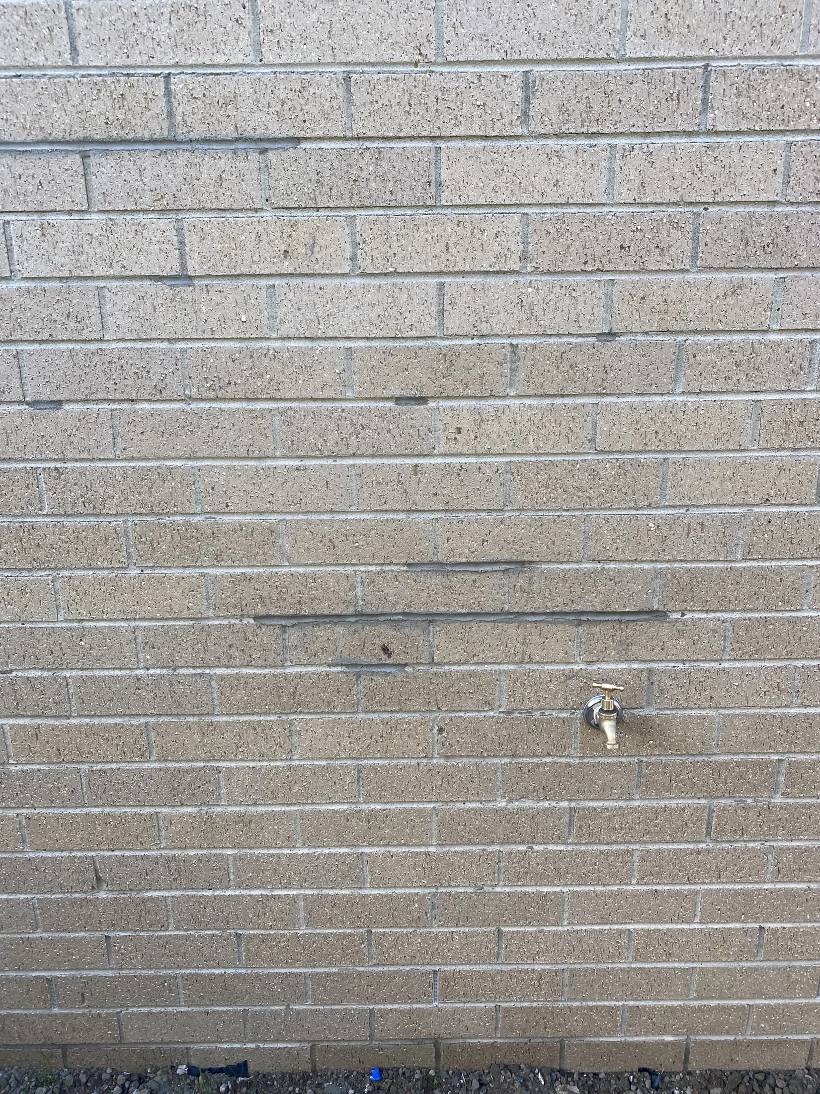 Tan brick wall with a silver outdoor faucet in the center. The ground is gravelly, and there's no visible text or notable patterns.