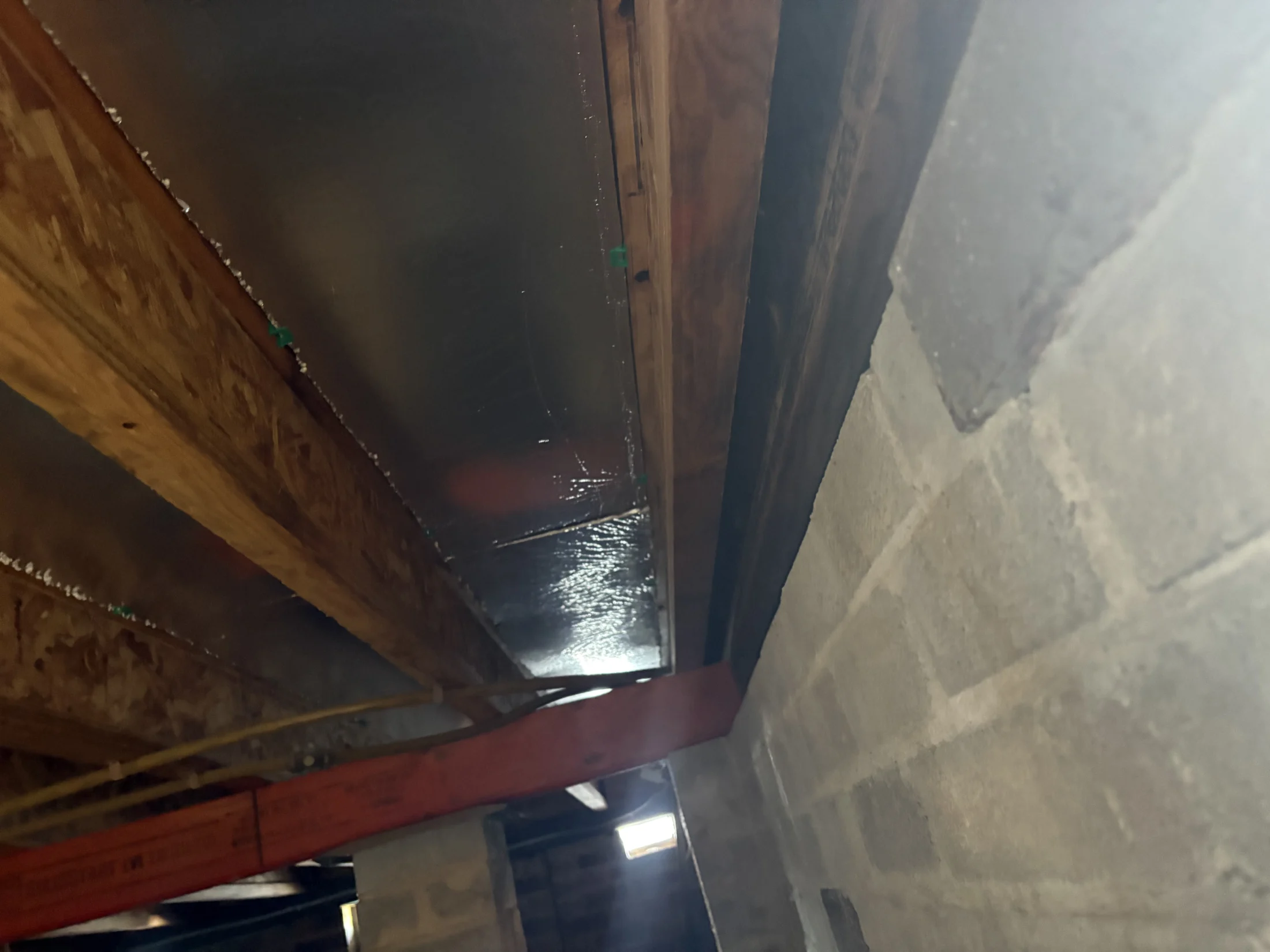 Wooden beams with insulation in a dimly lit basement. Concrete block wall on the right. Ceiling reflects light.