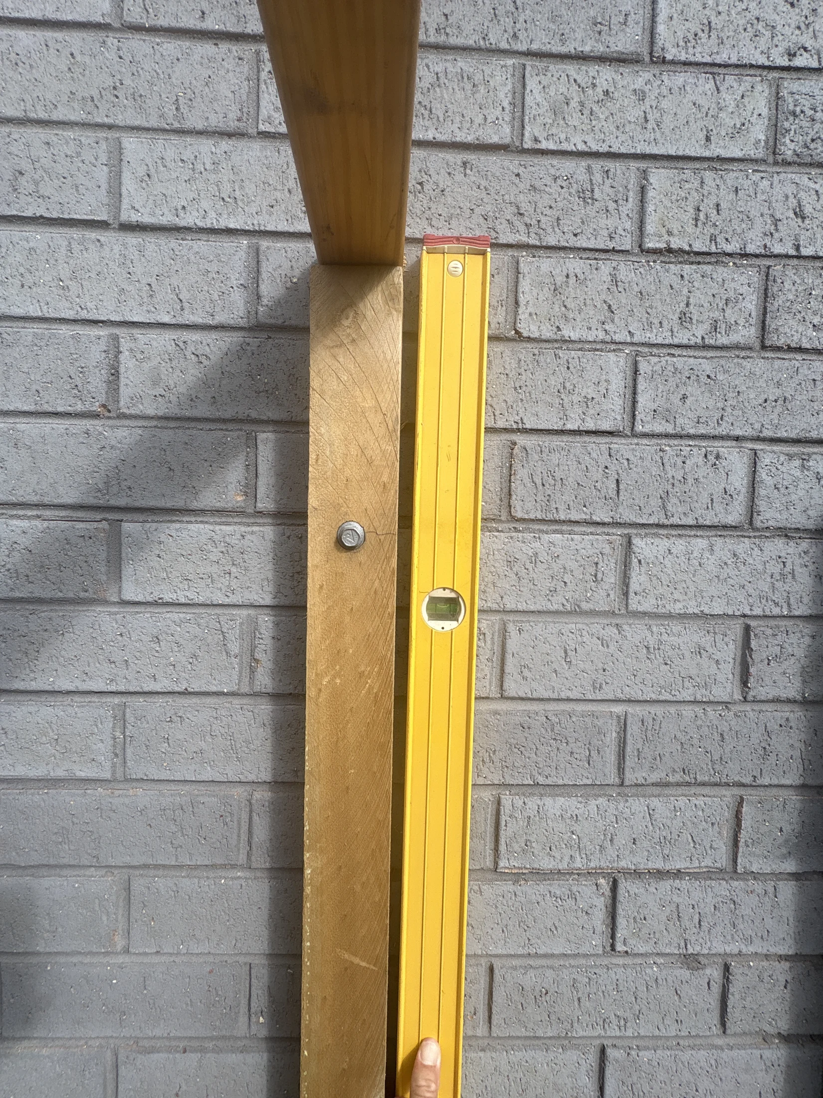 Yellow level held against a wooden beam, set on a gray brick wall. A hand supports the level at the bottom. Mood is focused.