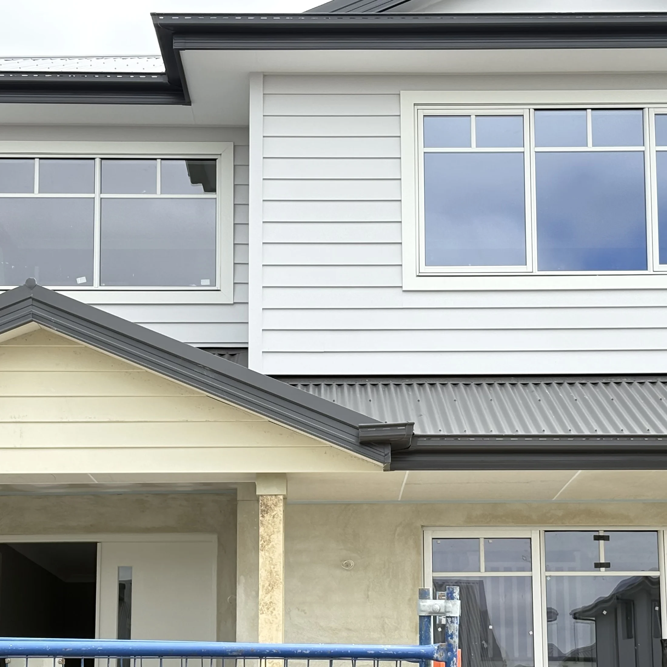 Two-story house exterior with light gray siding, large windows, and a metal roof. Blue sky reflects in windows, creating a calm mood.