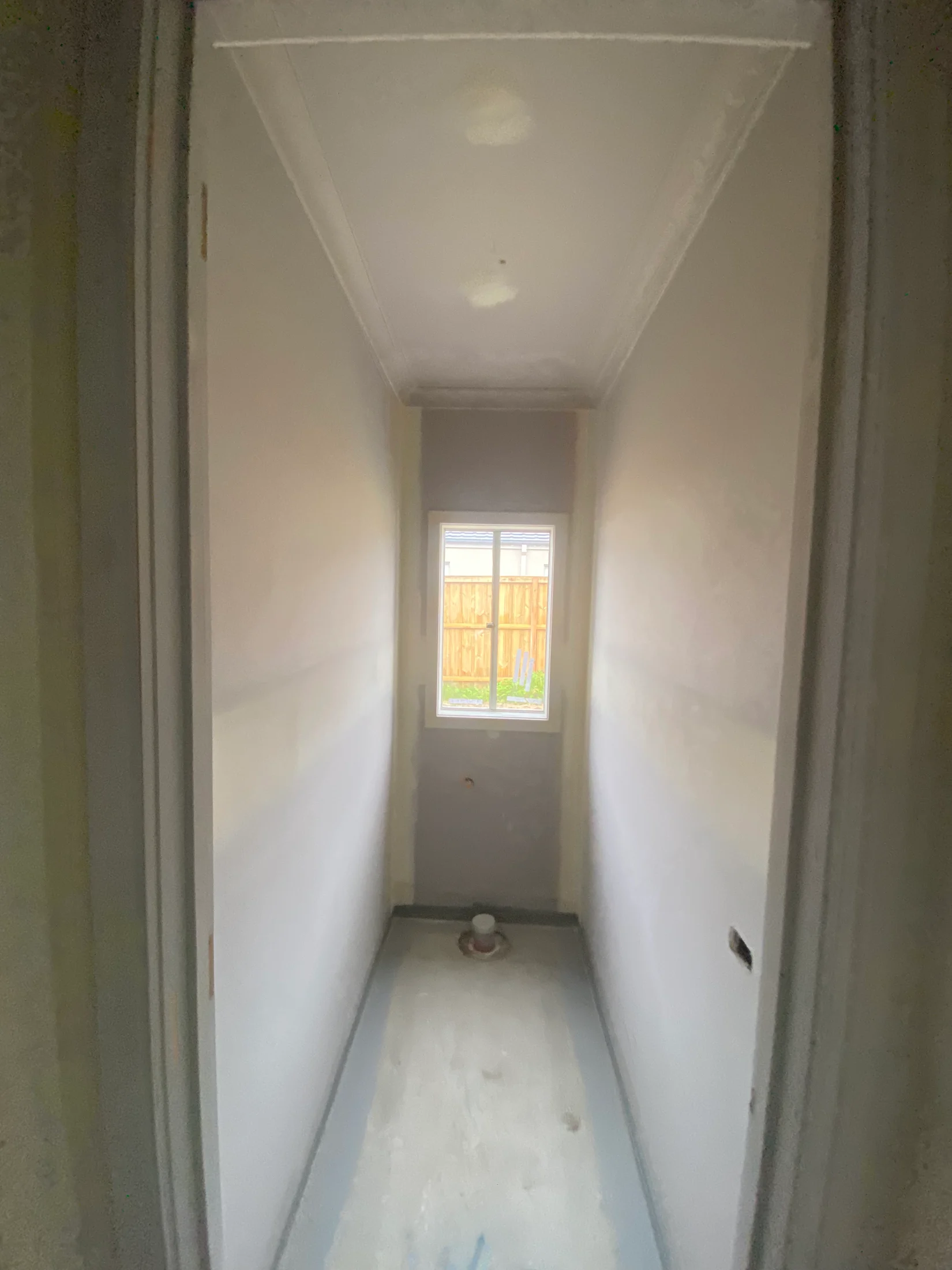 Narrow, empty room with light gray walls, small window at end. Ceiling has visible patches; floor is bare. Mood is minimalist and unfinished.