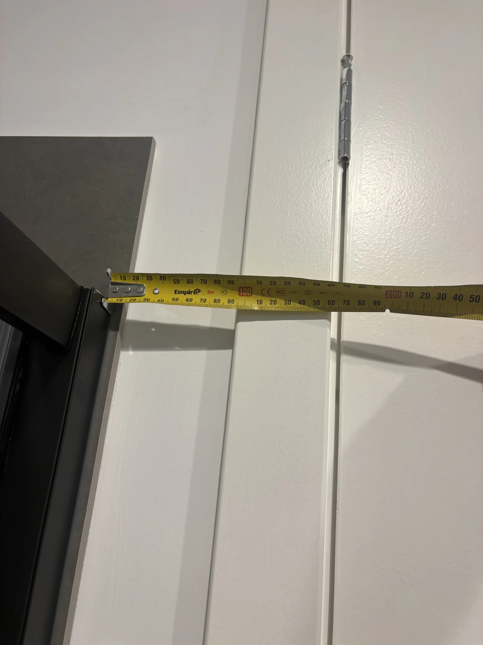 A yellow tape measure extended across a white door and dark metal frame, with measurements shown. Door hinge visible in the background.