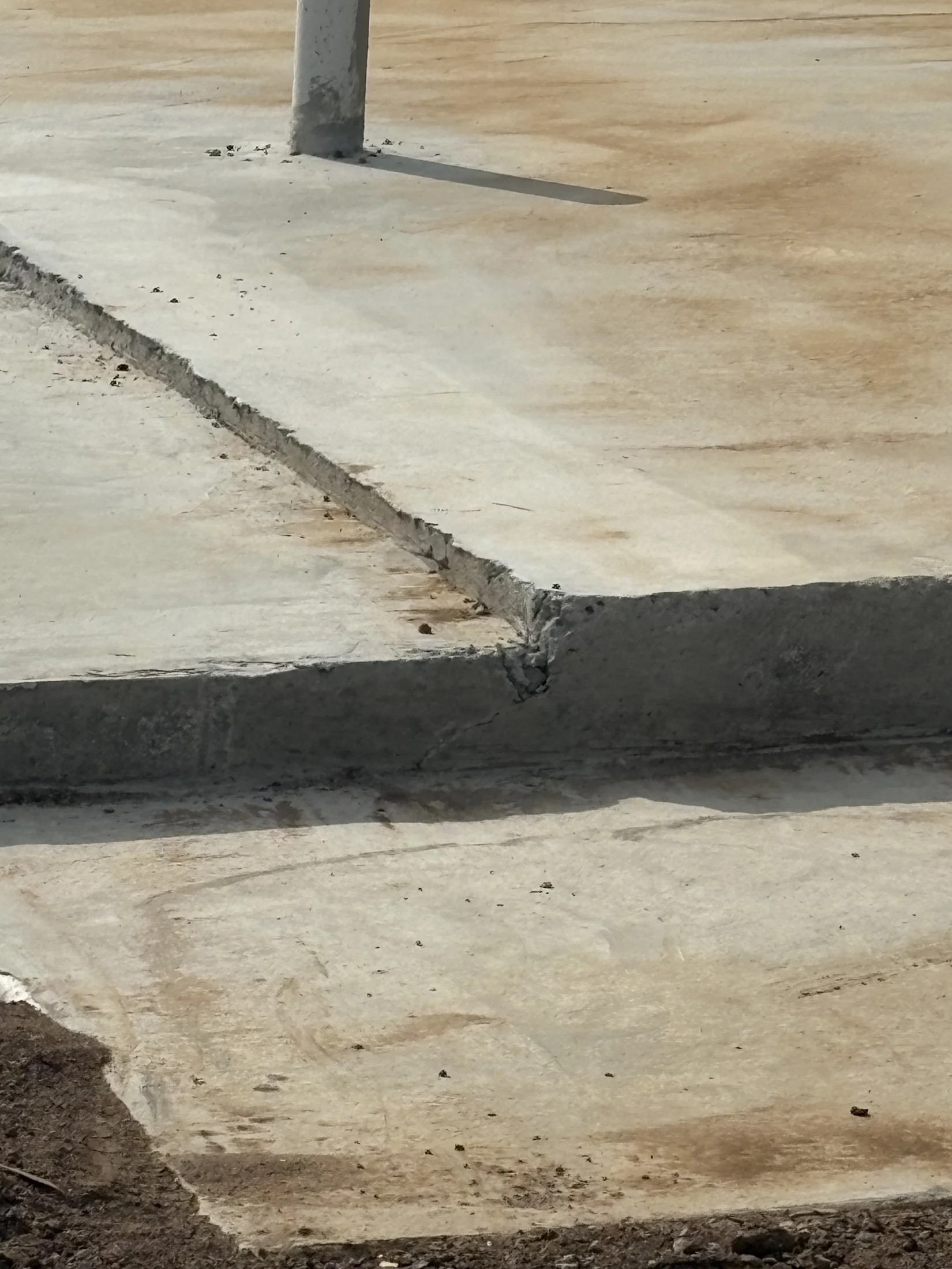 A concrete slab with a pole and a rough, uneven edge; cracks and dirt are visible on the surface, with a dry, dusty environment.
