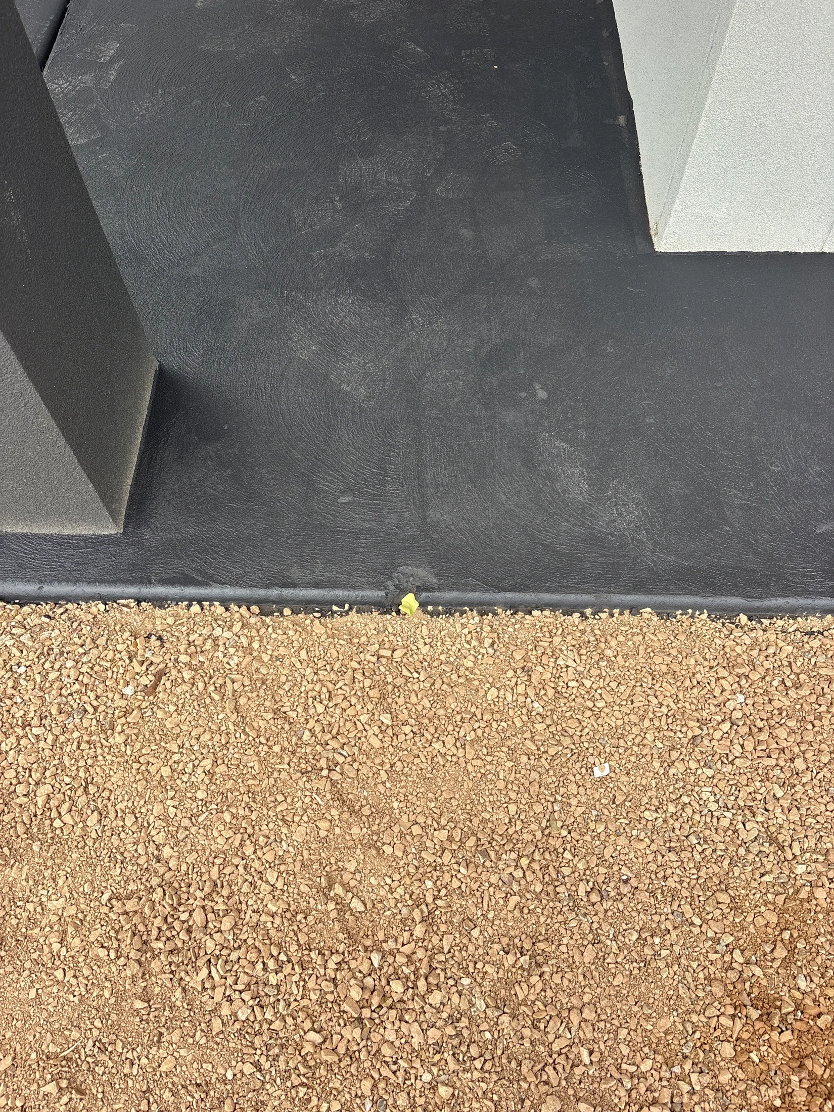 A black concrete surface meets a light brown gravel path. A small yellow object is visible between them. The scene is calm and minimalistic.