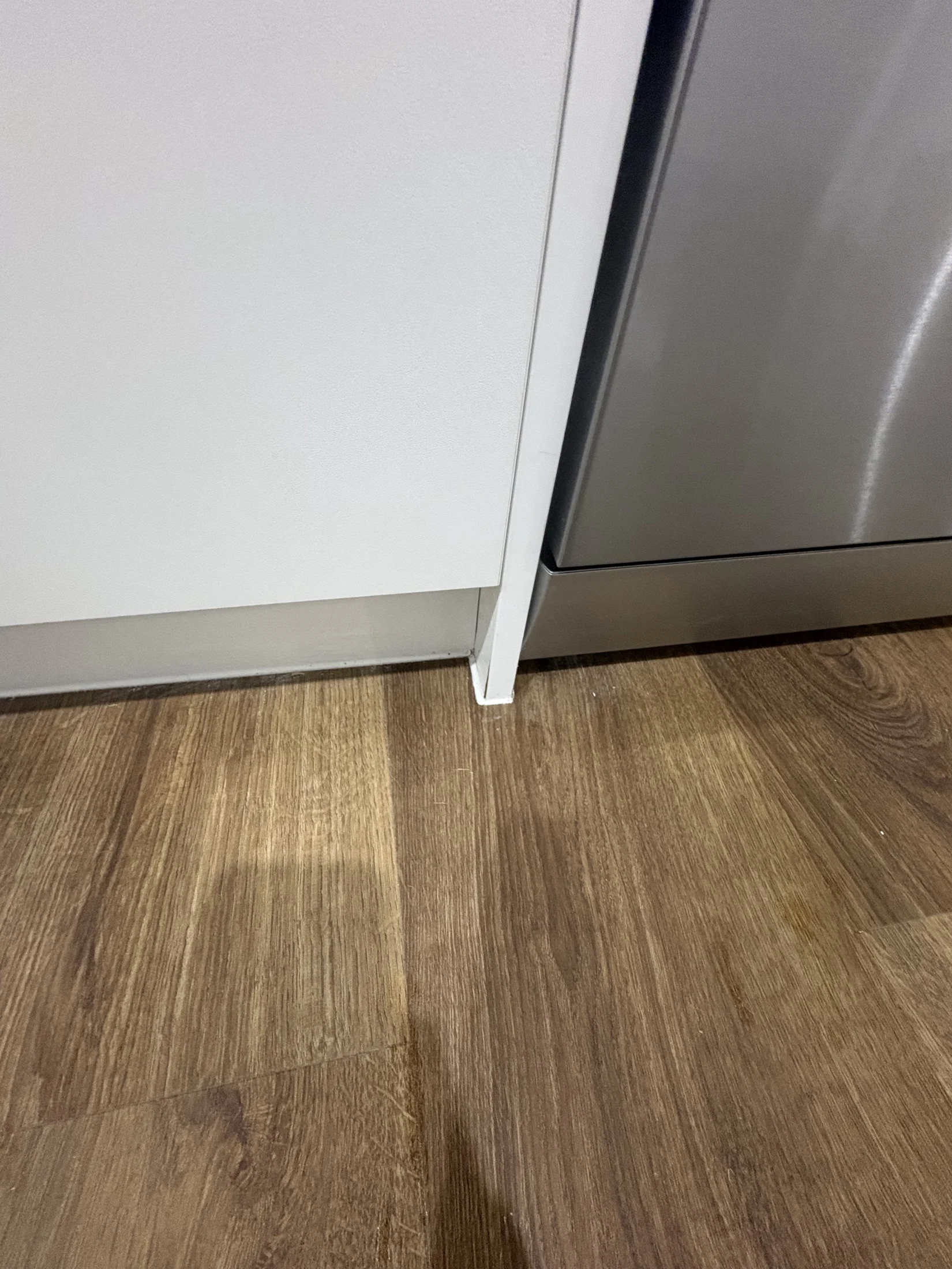 Wooden floor meets a white cabinet and stainless steel appliance in a kitchen setting. Light from above casts a shadow on the floor.