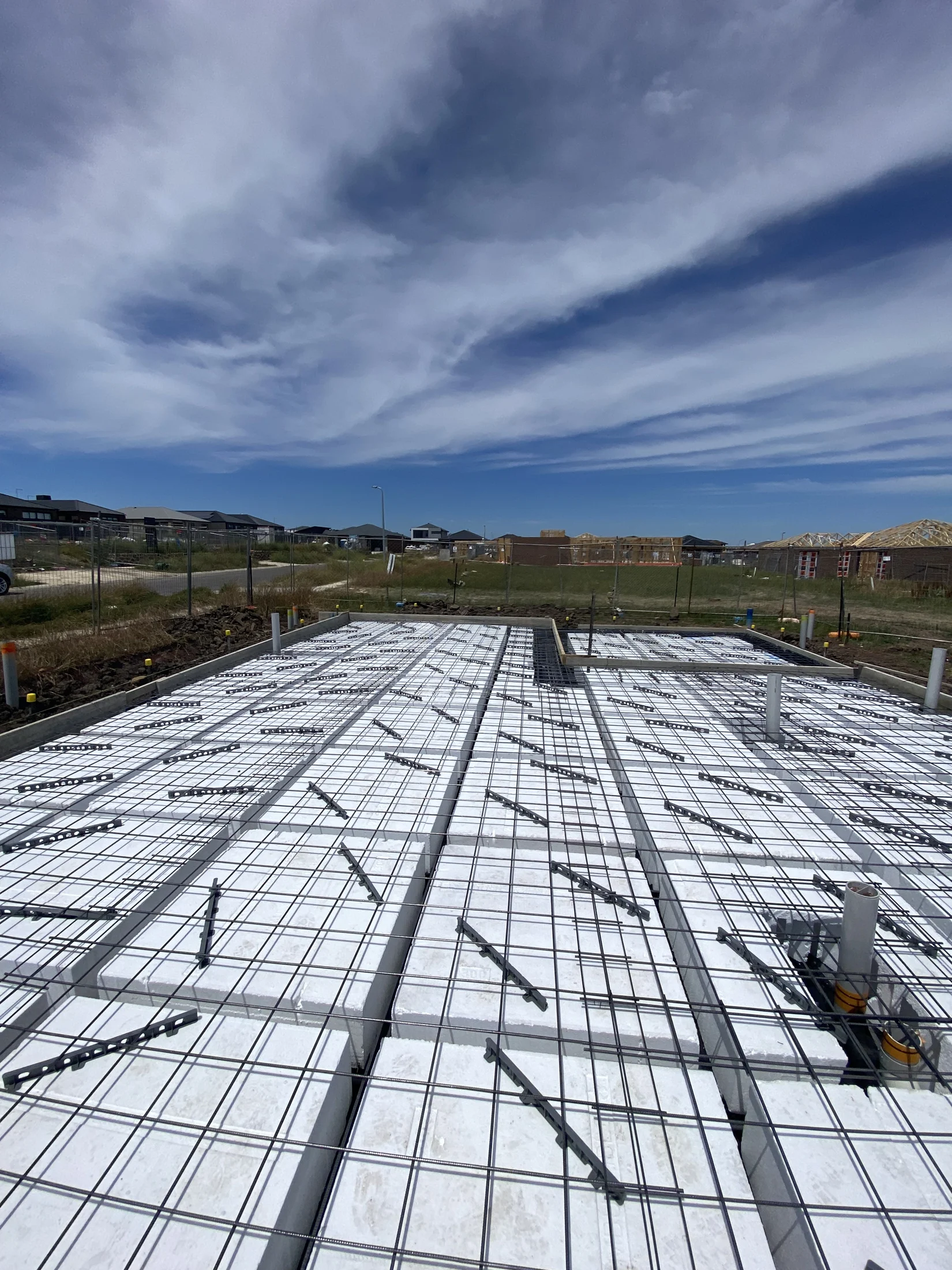 Concrete slab with reinforcing bars on a construction site under a blue sky with clouds. Partially built houses visible in the background.