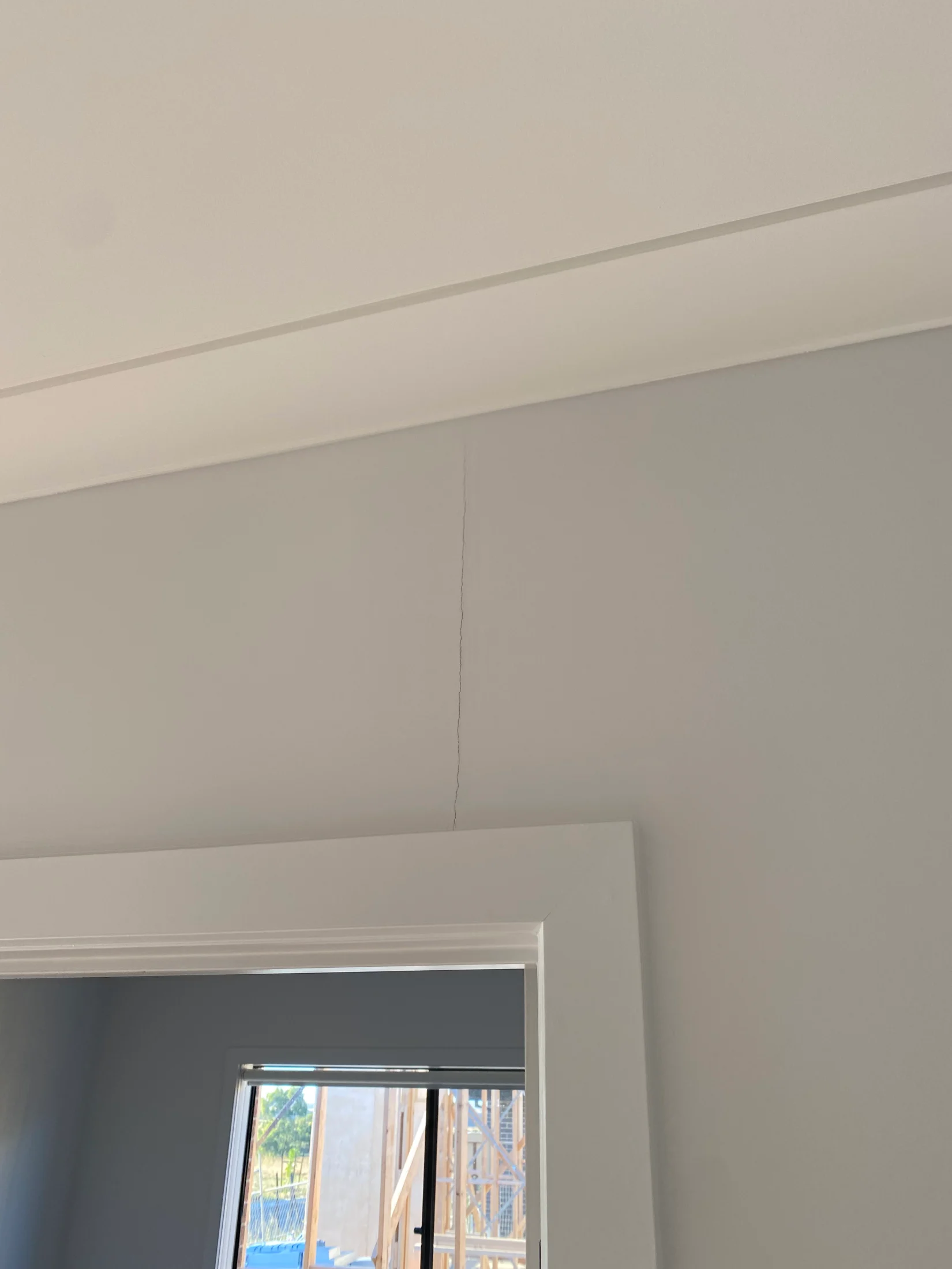 Crack above a white door frame on a light gray wall. Sunlit window shows construction scene outside, creating a slightly worn atmosphere.