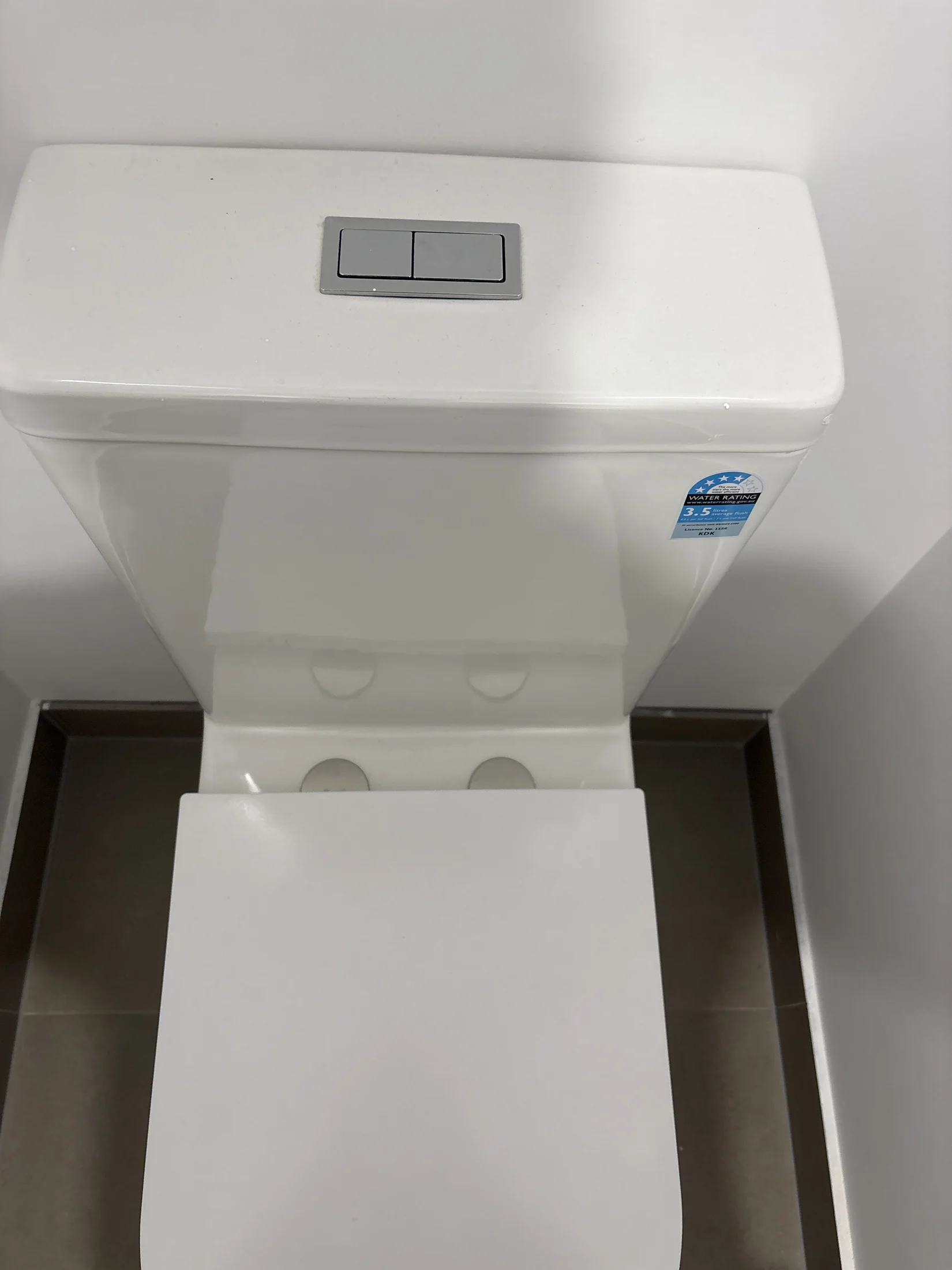 White toilet with dual flush buttons, viewed from above. Water-saving sticker visible. Clean, minimal bathroom setting.