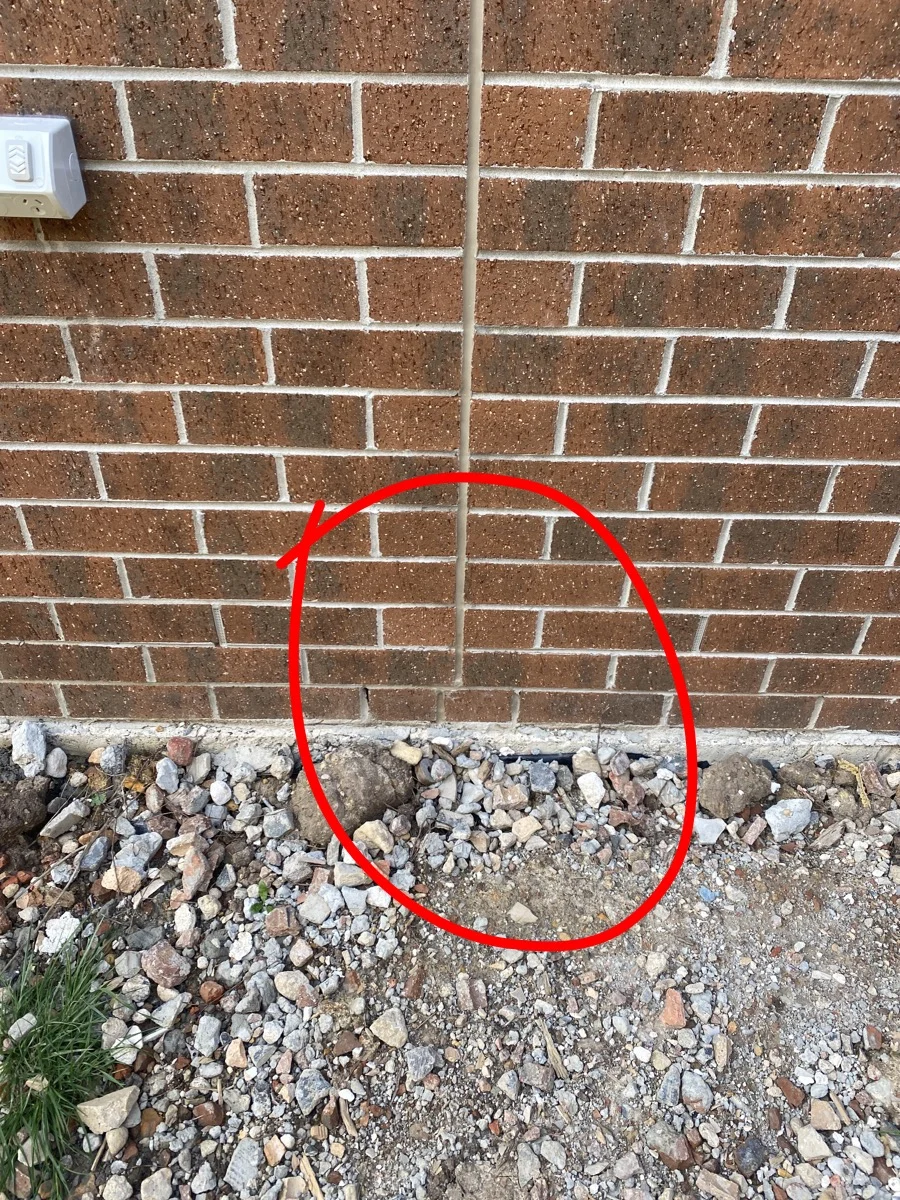 Brick wall with vertical crack highlighted by a red circle. Loose gravel and rocks on the ground. An electrical outlet is visible.