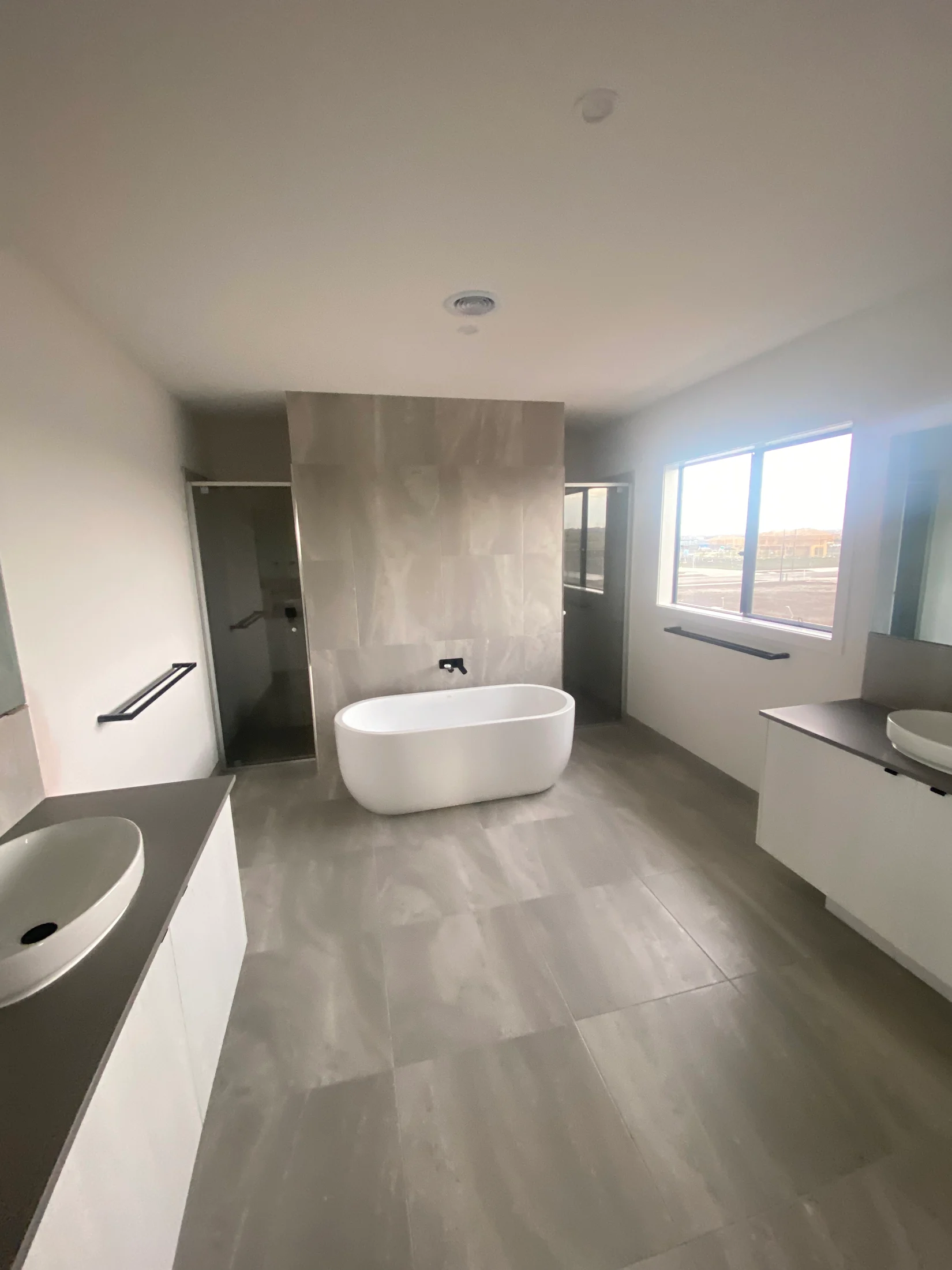 Spacious bathroom with a freestanding white tub, dual sinks, and large window. Soft gray tiles and minimalist design create a serene atmosphere.