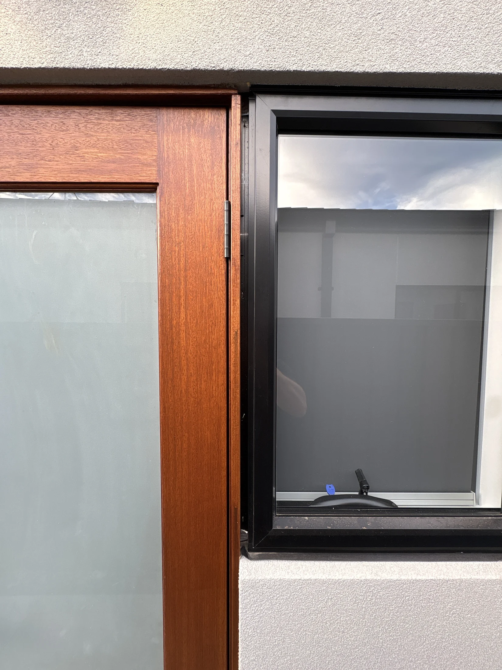Wooden door and black-framed window close-up. The setting has a modern, minimalistic exterior. Sky reflection visible in the window.