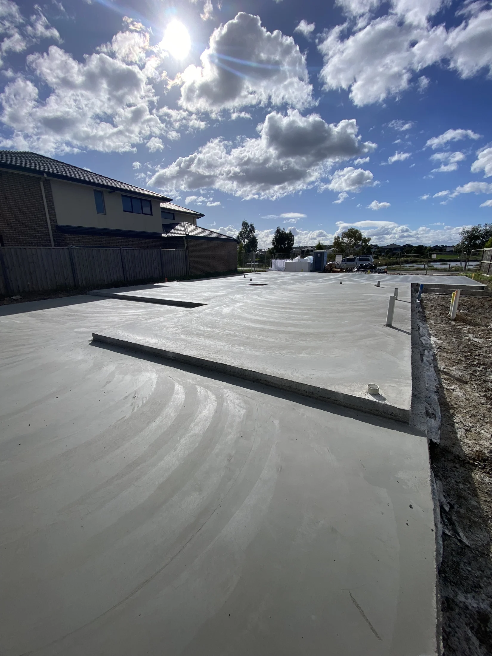 Fresh concrete slab in a sunny suburban area with a brick house and cloudy blue sky. Shadows cast on the smooth surface.