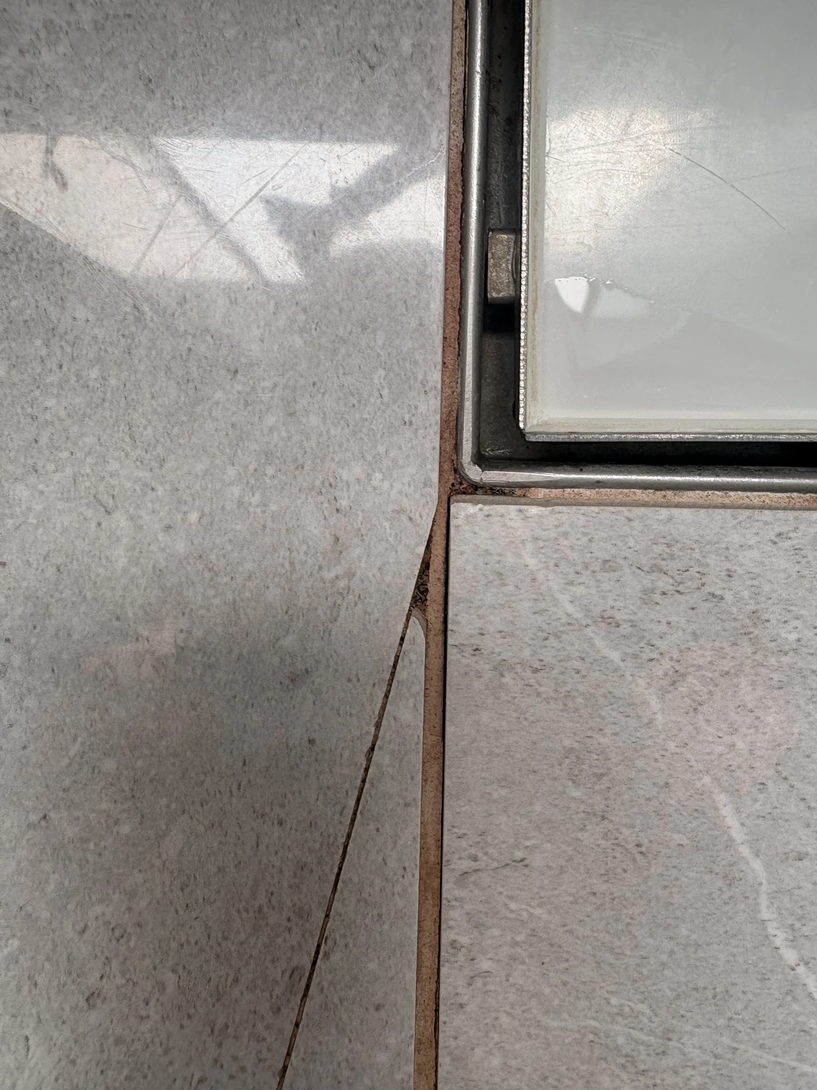 Close-up of light gray tiles with a small gap in between. Slight reflections are visible on the surface, and a shadow casts a subtle pattern.
