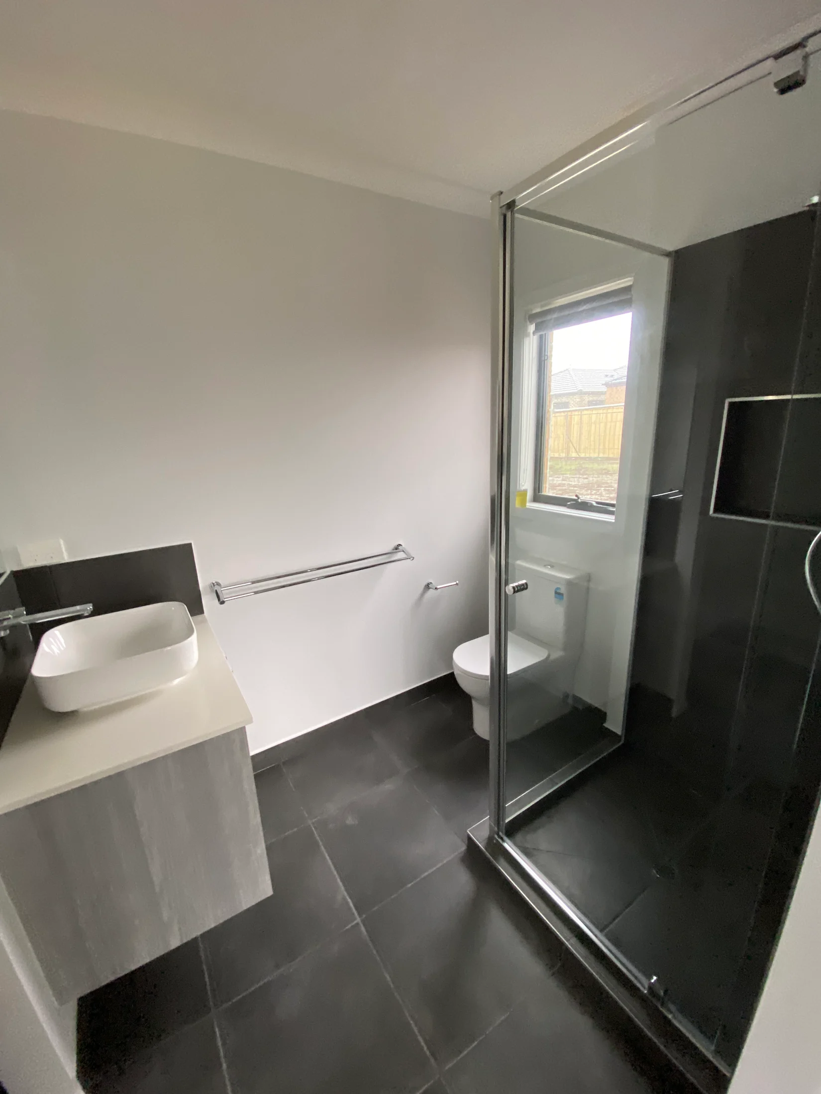 Modern bathroom with black tiles, a glass shower, white vanity and sink. A window lets in light, creating a clean, minimalistic feel.