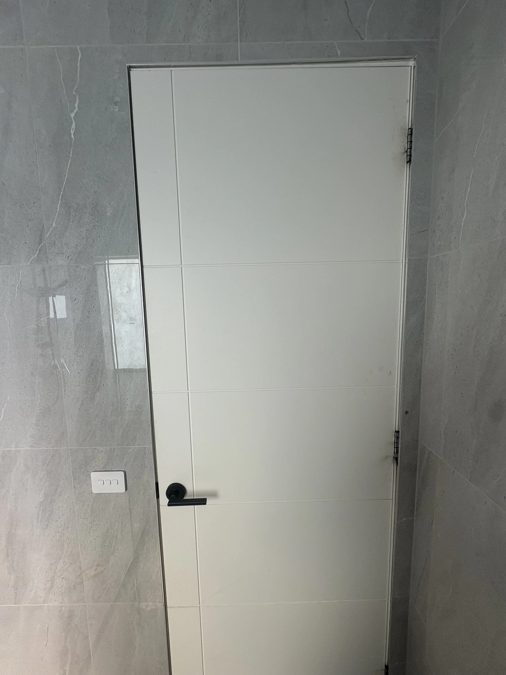 White door with black handle in a gray tiled room. Slight discoloration near the door hinges. Light switch on the wall beside the door.