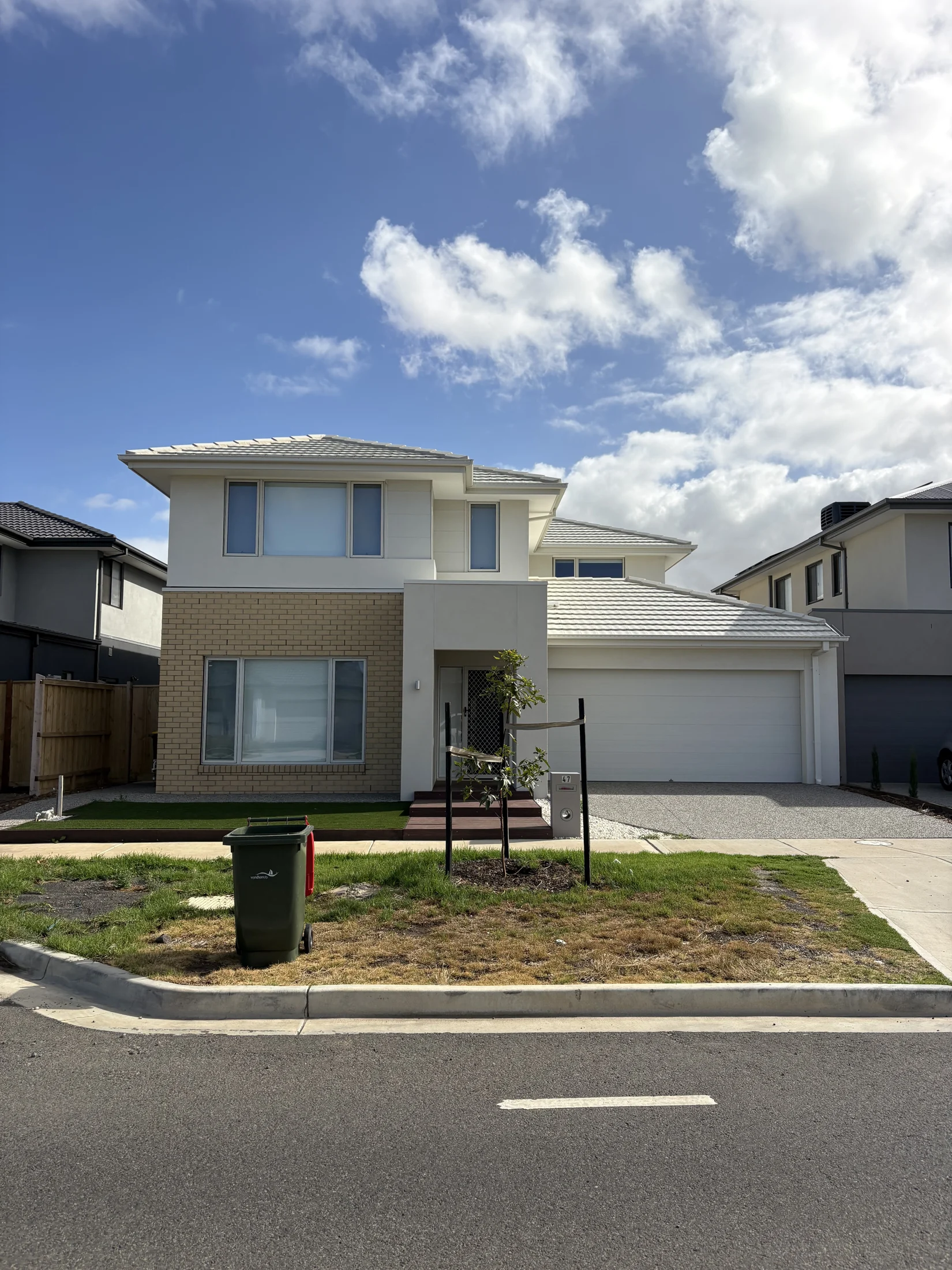Modern two-story house with a beige brick facade, green lawn, and a small tree in front. Blue sky and clouds in the background.