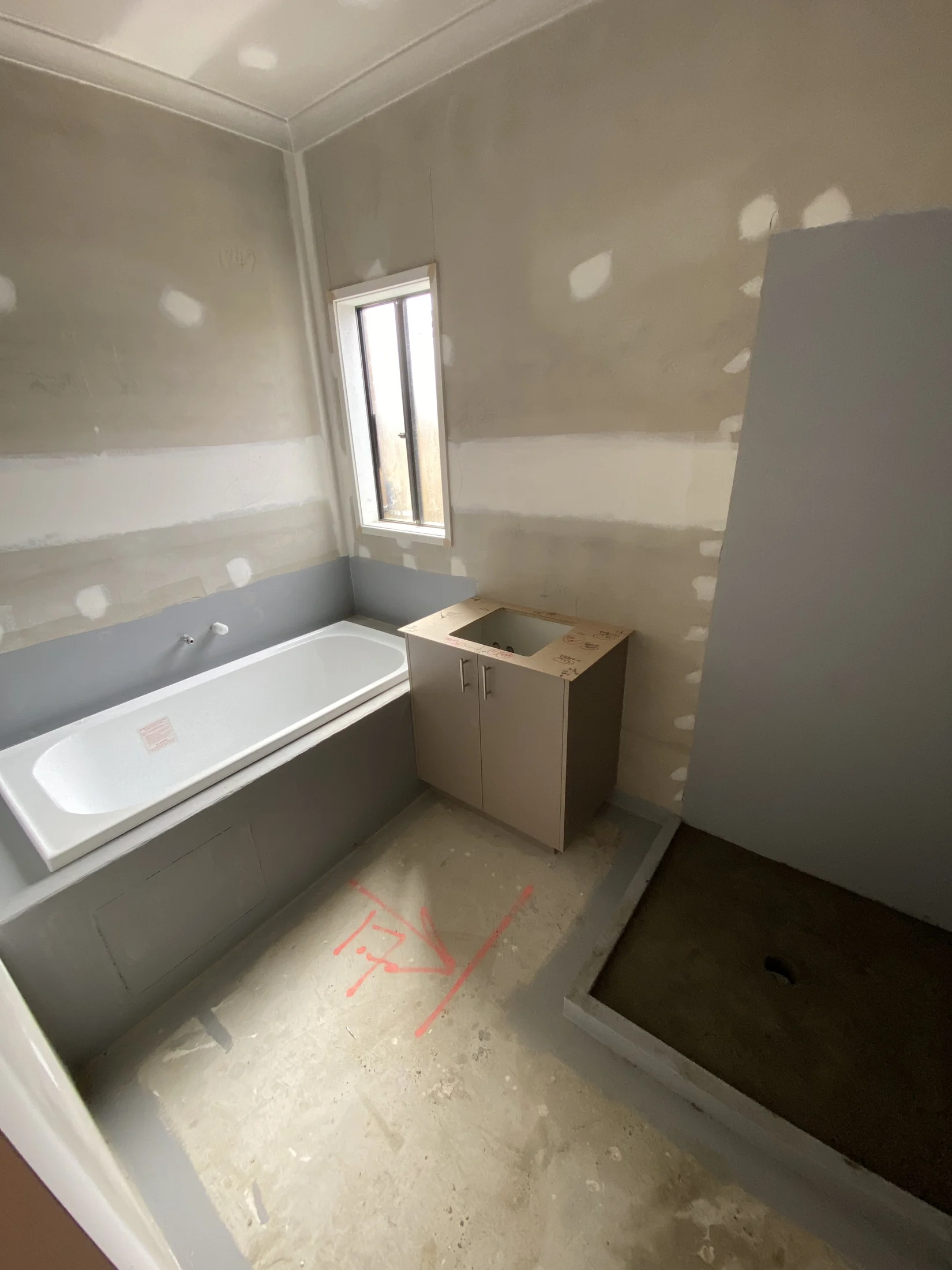 Unfinished bathroom with a tub, cabinet, and shower area. Gray walls with visible patches; red markings on the floor. Mood is neutral.