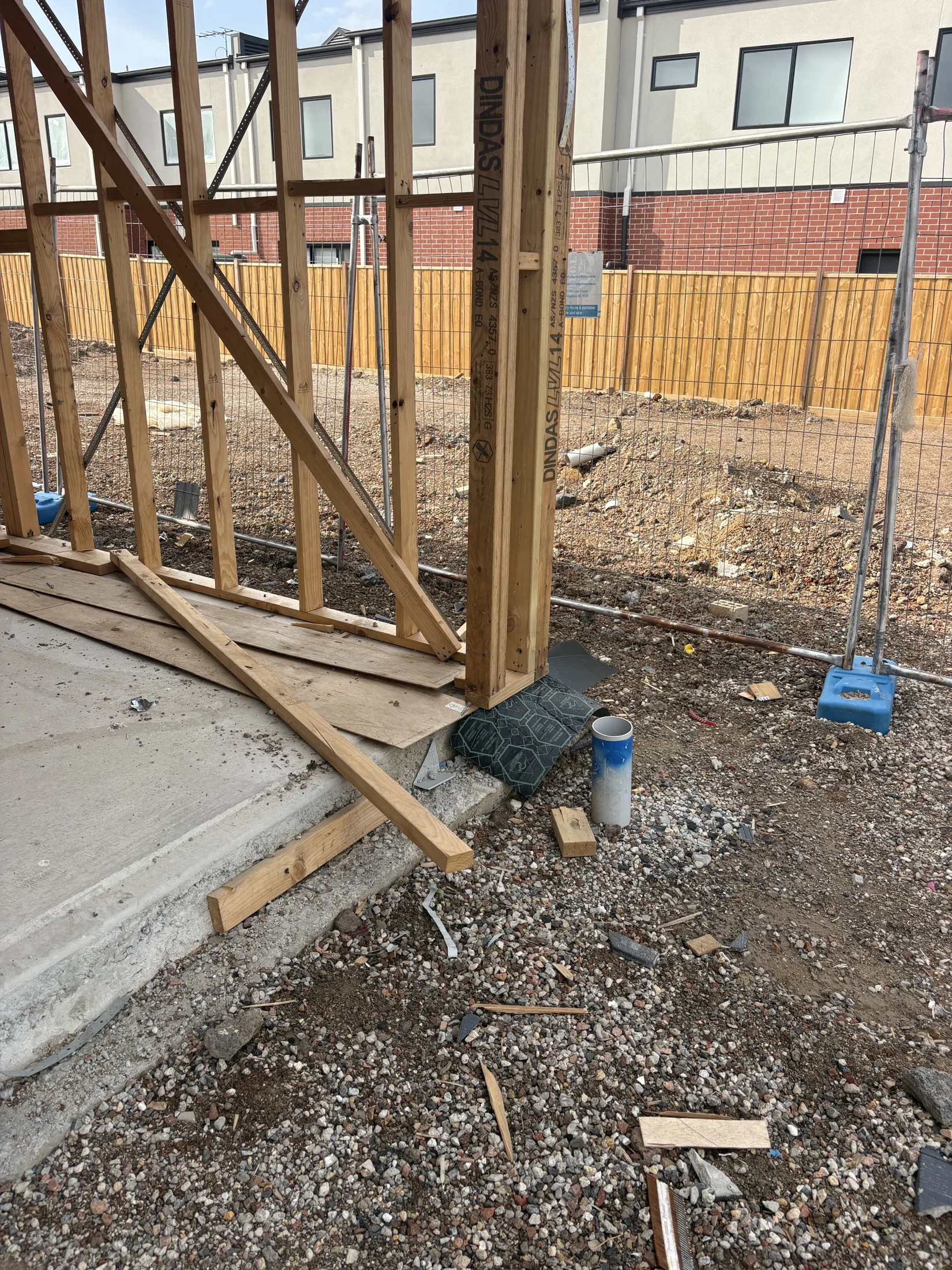 Wooden frame construction site with scattered debris and gravel ground. Nearby are a brick and wooden fence, and adjacent buildings.