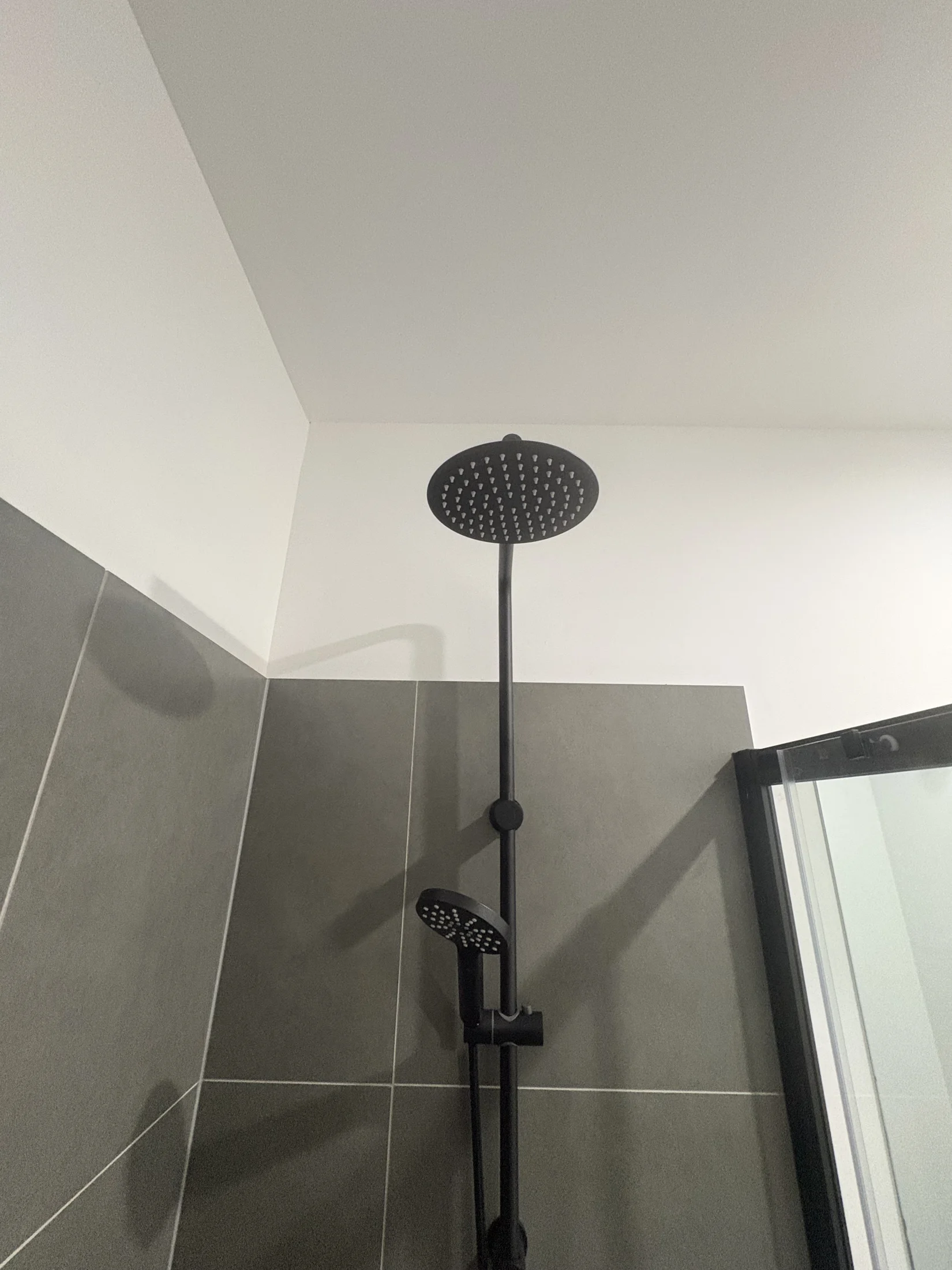 Black showerhead in a modern bathroom with grey tiled walls and a glass shower door, creating a sleek and minimalist look.