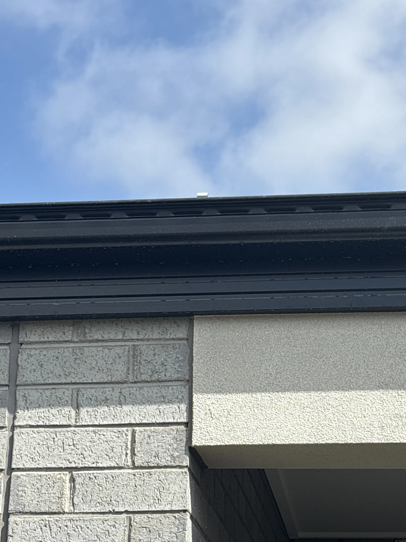 Building corner with gray brick wall and beige accents under a blue sky with clouds. Black gutter at roof edge, no action or text visible.