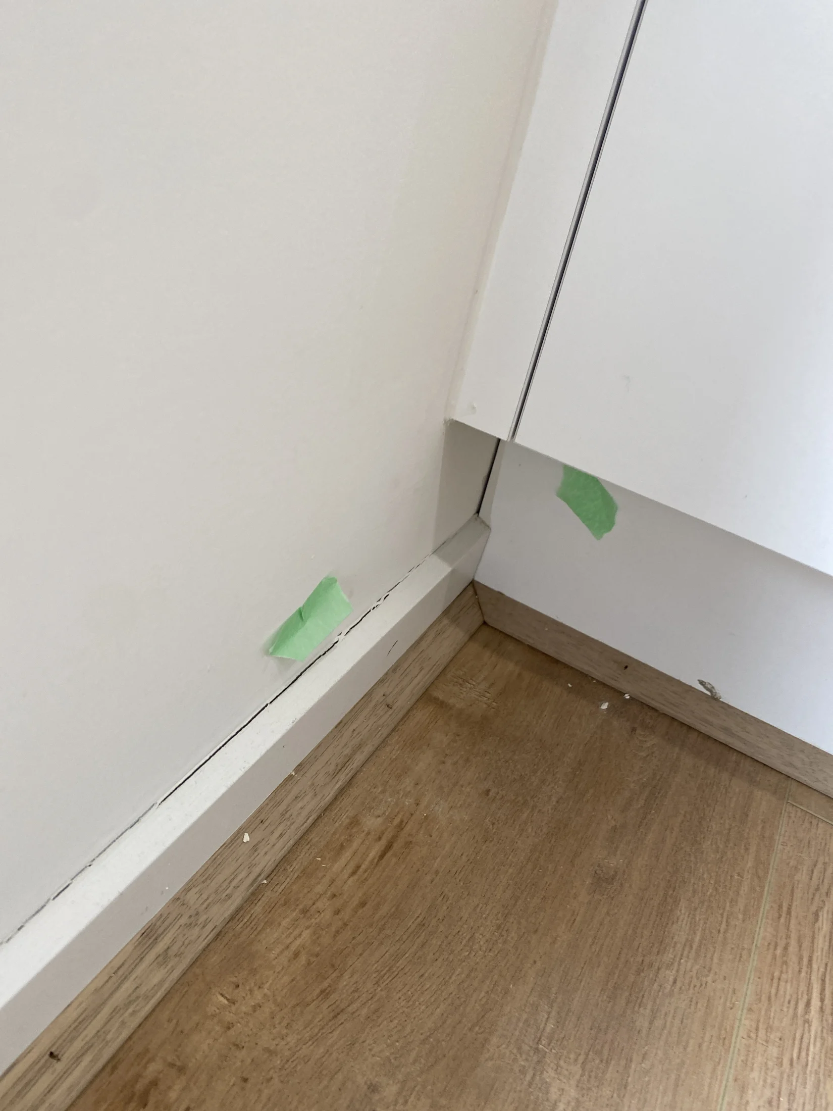 Cracked white wall corner with green tape marking the damage. Light wood floor and baseboard contrast the stark wall. Minimalist setting.