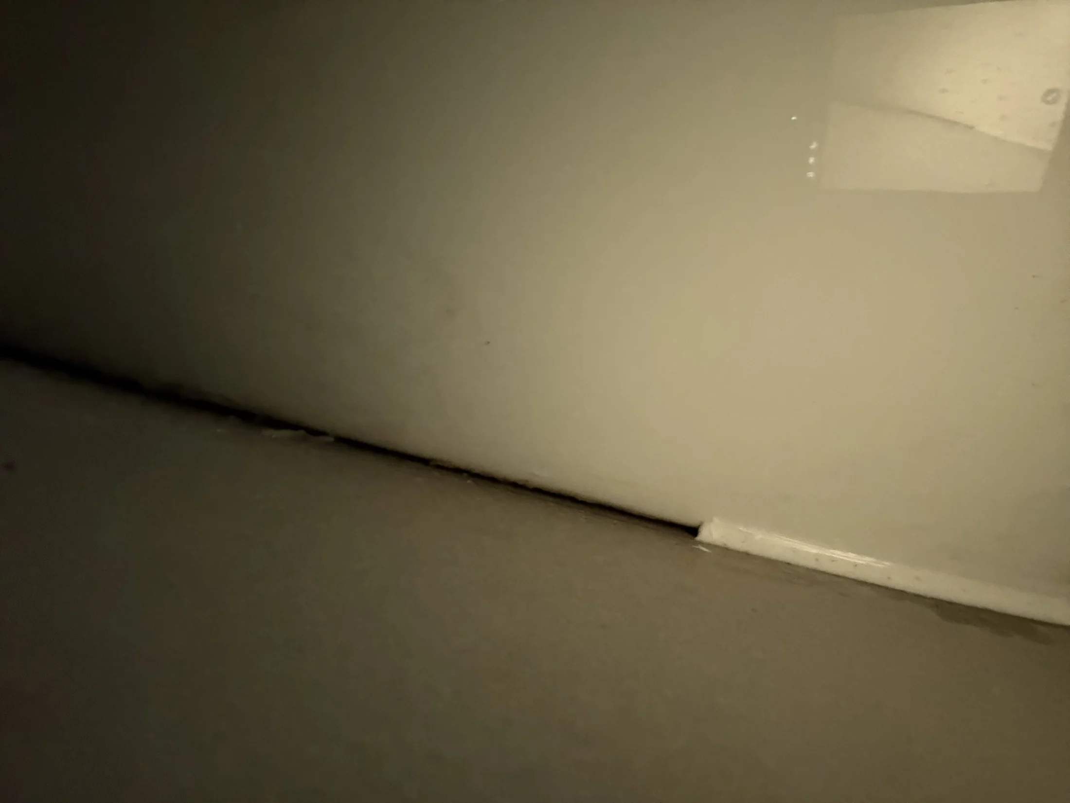 Dimly lit close-up of a beige wall and floor corner, with faint reflections. The setting appears empty and quiet, creating a subdued mood.