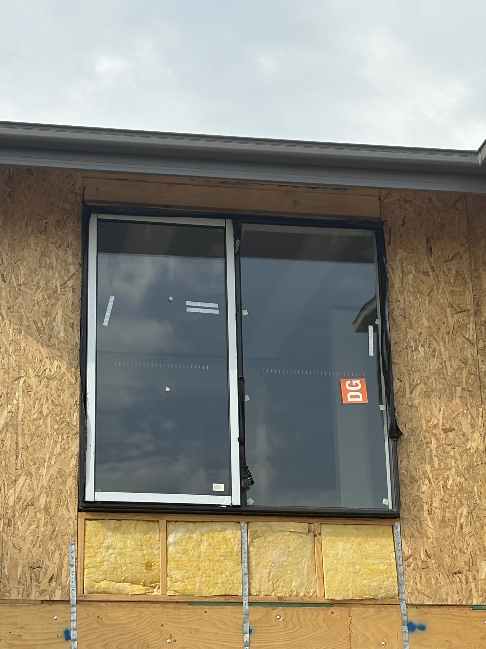 Two large windows on an unfinished wall with wood paneling and yellow insulation. A label "DG" is visible on one window. Cloudy sky above.