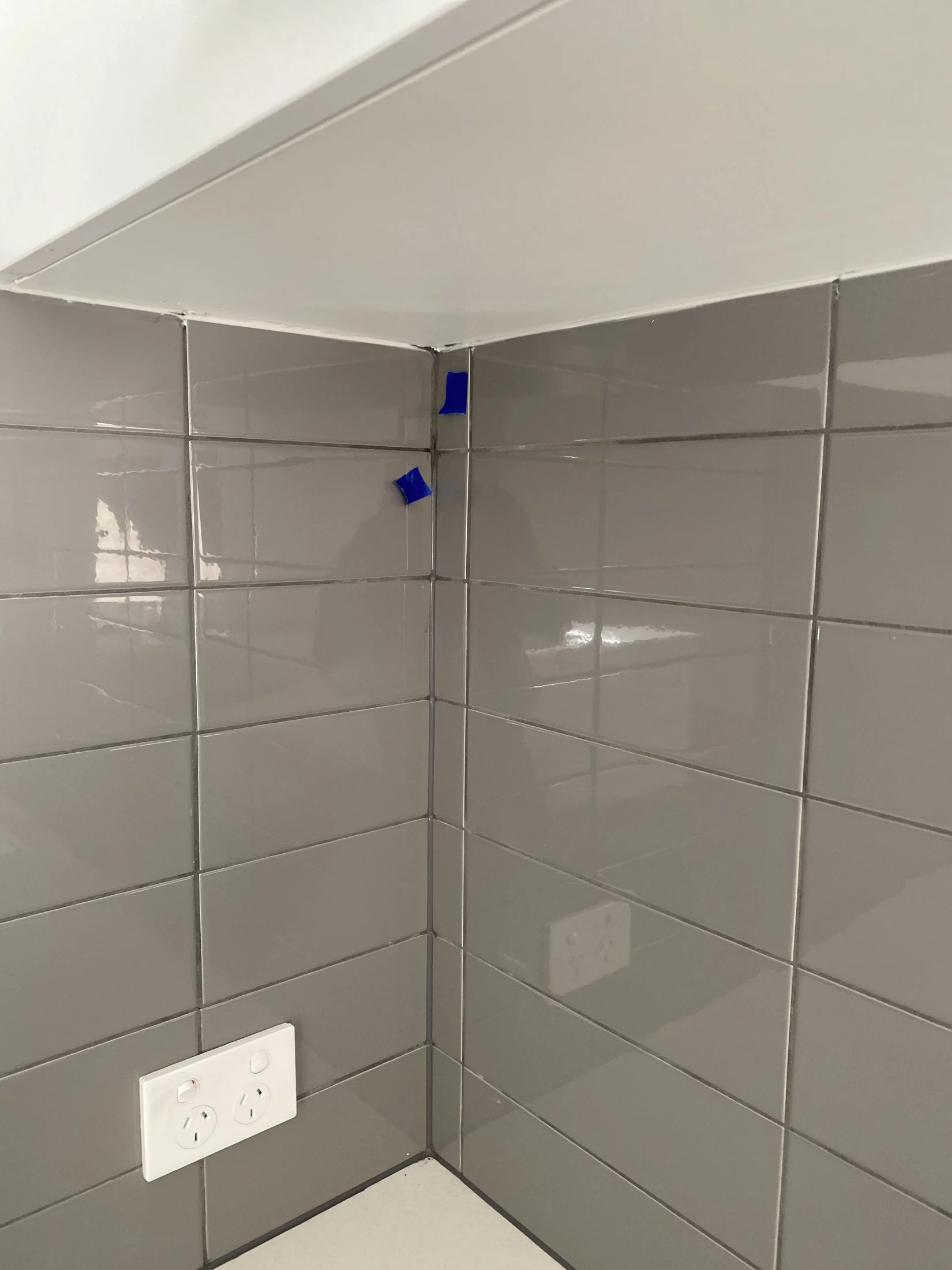 Gray tiled kitchen corner with blue tape markings, a white countertop, and a double power outlet. Bright lighting and modern feel.