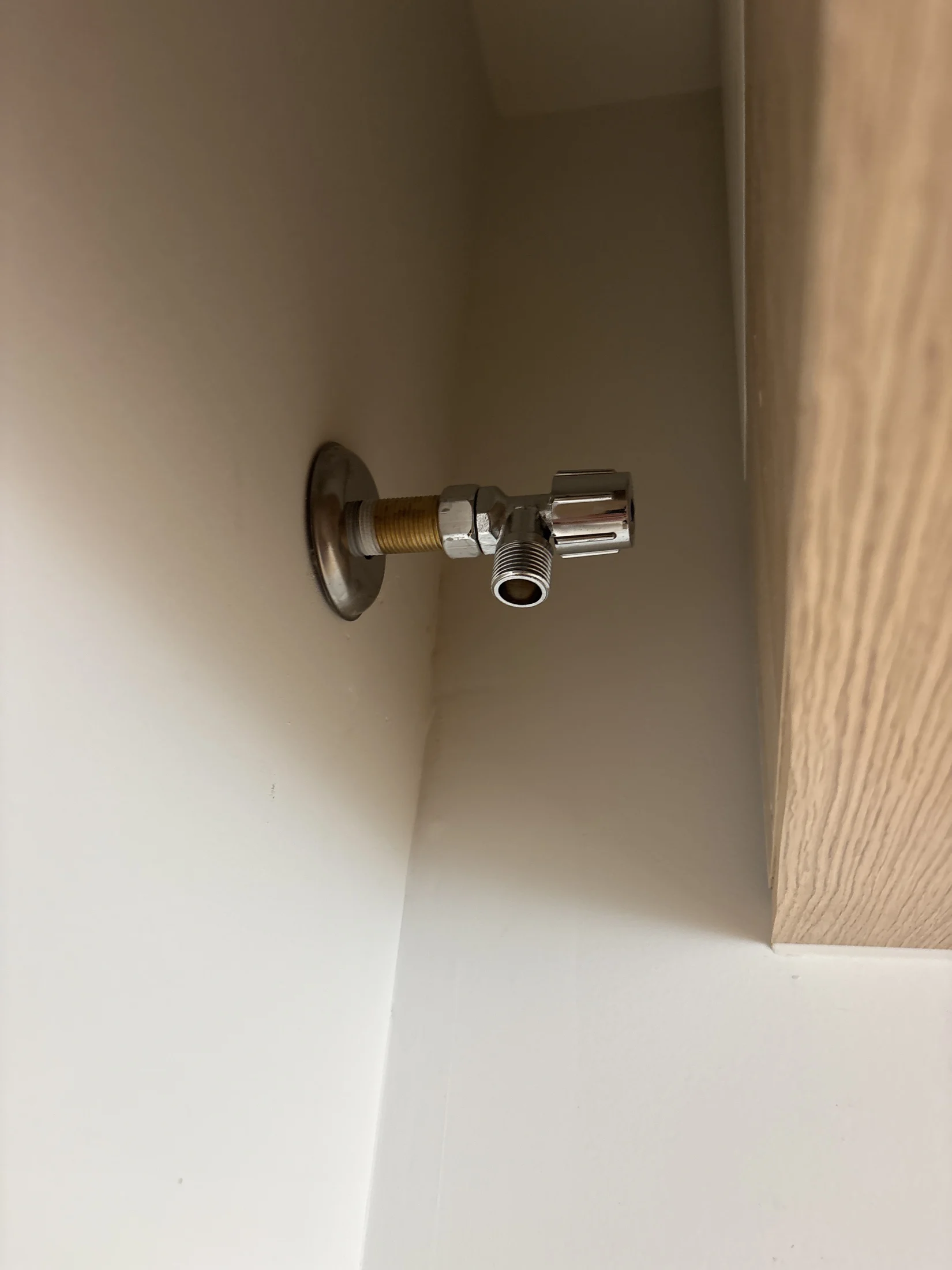 Close-up of a shiny metal valve with yellow threading on a white wall. Wooden surface visible on the right. No text or notable action.