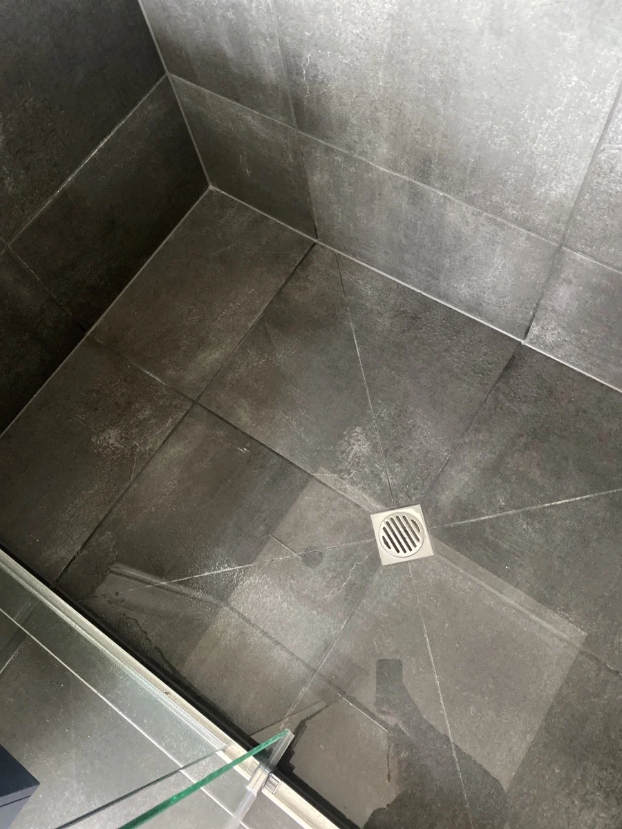 Shower floor with dark gray tiles, centered drain, and glass pane in foreground. The tiles have a subtle sheen, creating a sleek look.