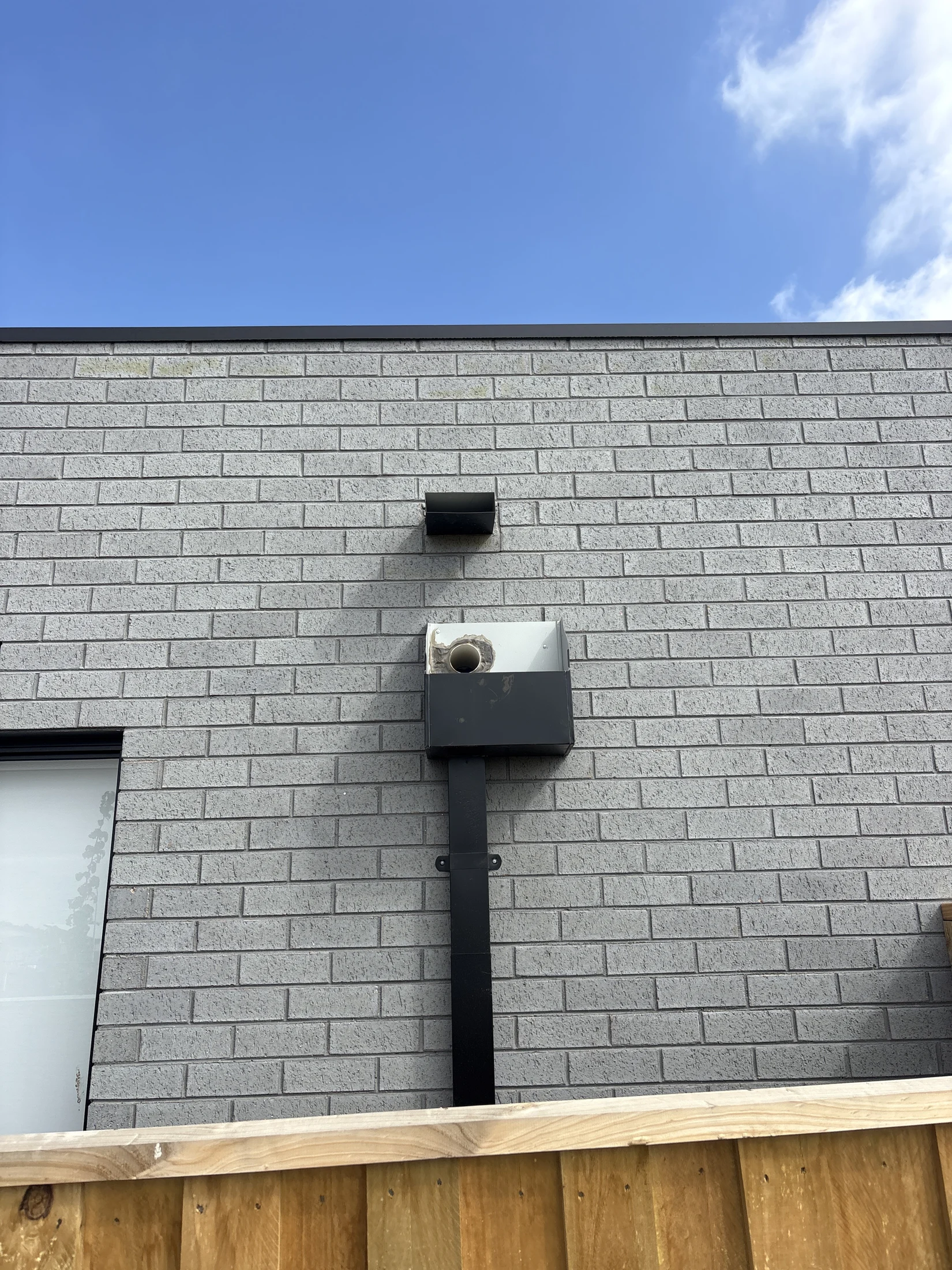 Gray brick wall with a vent and a camera mounted on a black pole. Clear blue sky above and wooden fence at the bottom.
