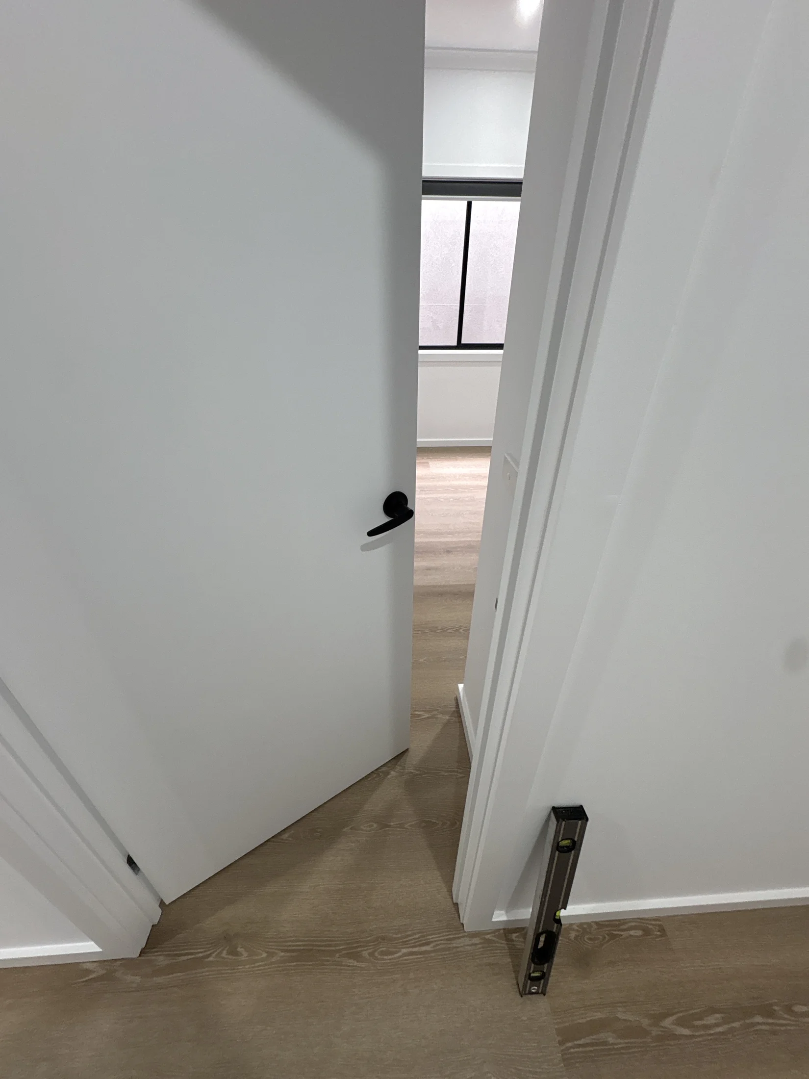 A partially open white door with a black handle, revealing a room with wood flooring. A level tool is on the floor, suggesting measurement.