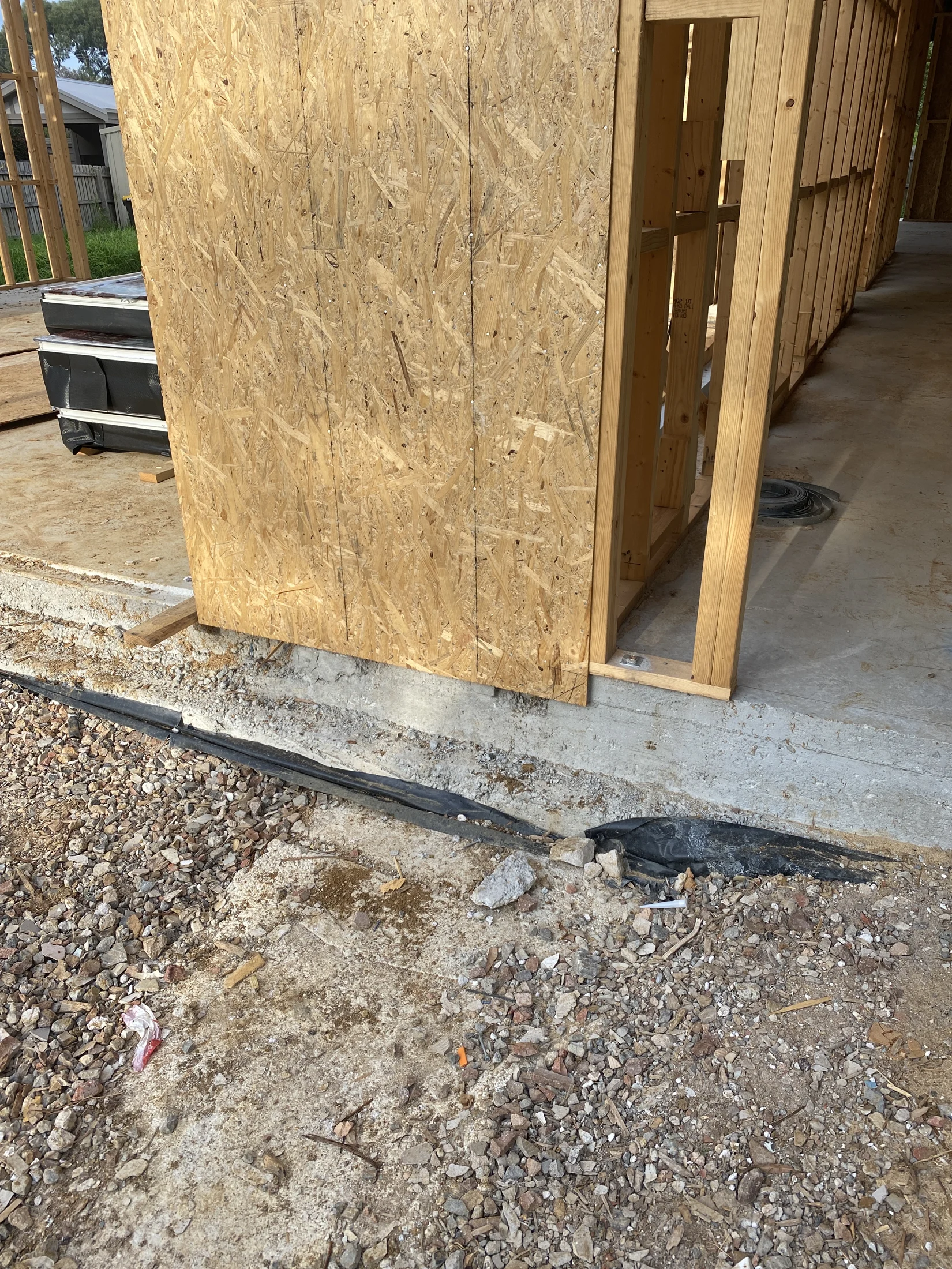 Wooden structure under construction with OSB panels, surrounded by gravel and dirt. A black tarp is partially visible on the ground.