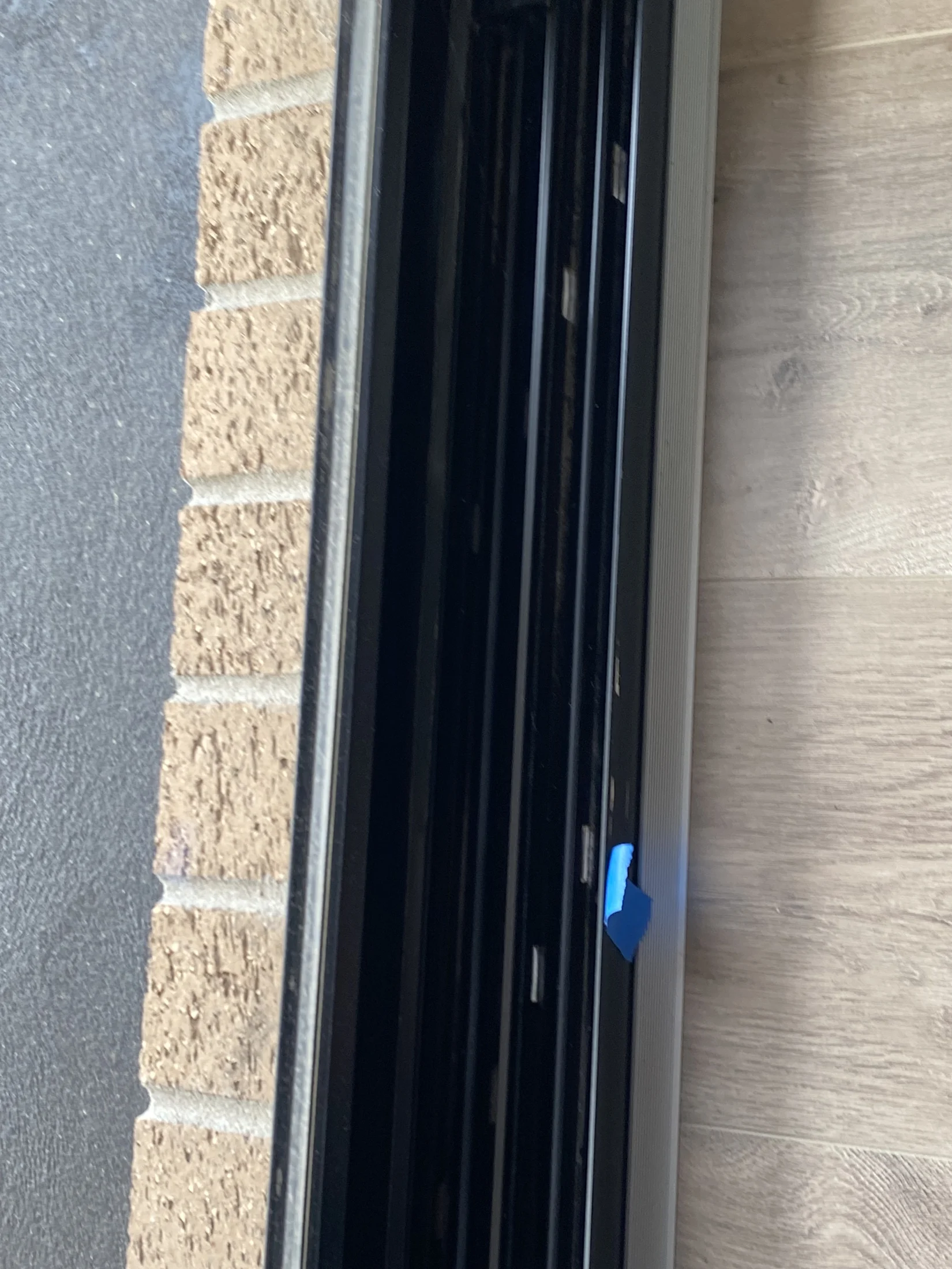 A brick wall meets a black door frame with a piece of blue tape on it. The floor is wooden. No text or people present.