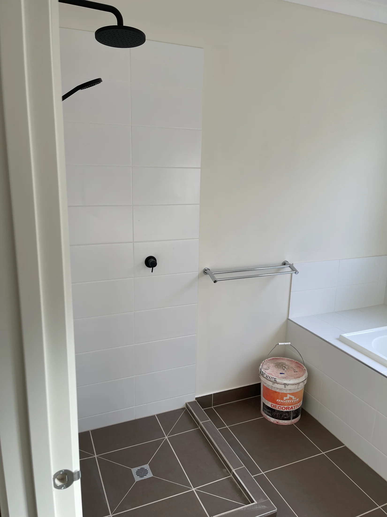 Modern bathroom with dark tiles, white tiled walls, a black ceiling shower, and a silver towel rack. A paint bucket sits near the tub.