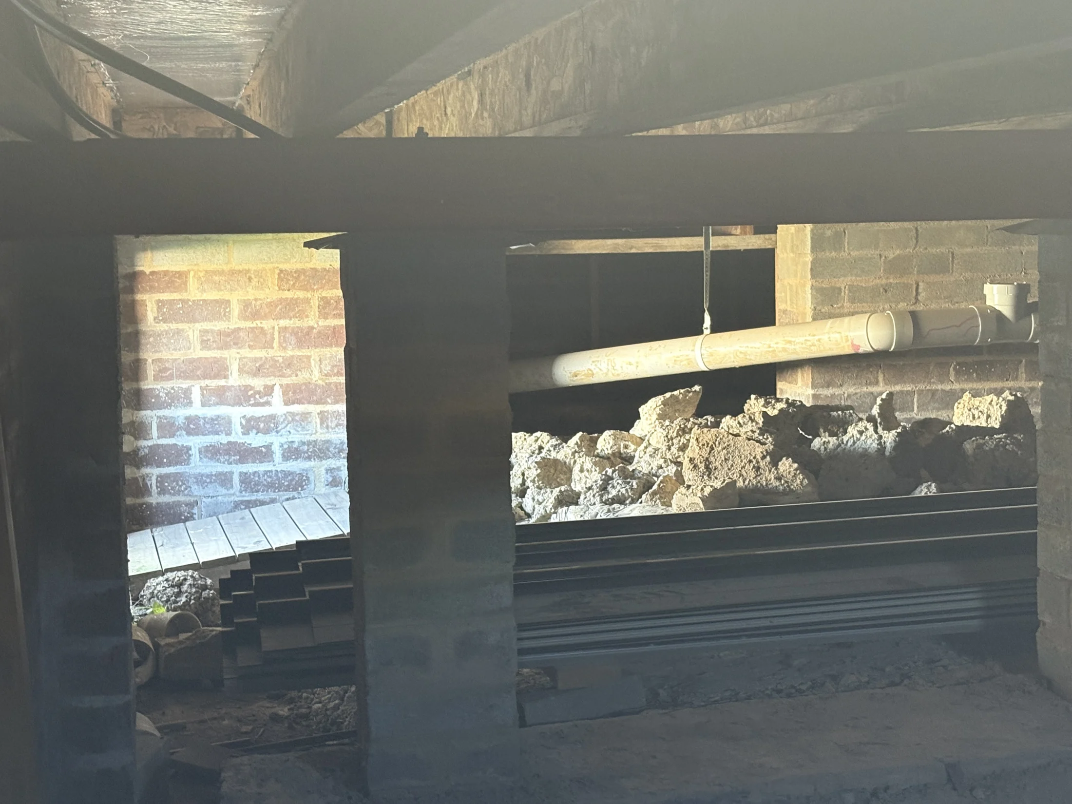 Under a house, a crawl space with brick walls and metal beams. A white pipe runs above piles of debris, lit by soft sunlight.