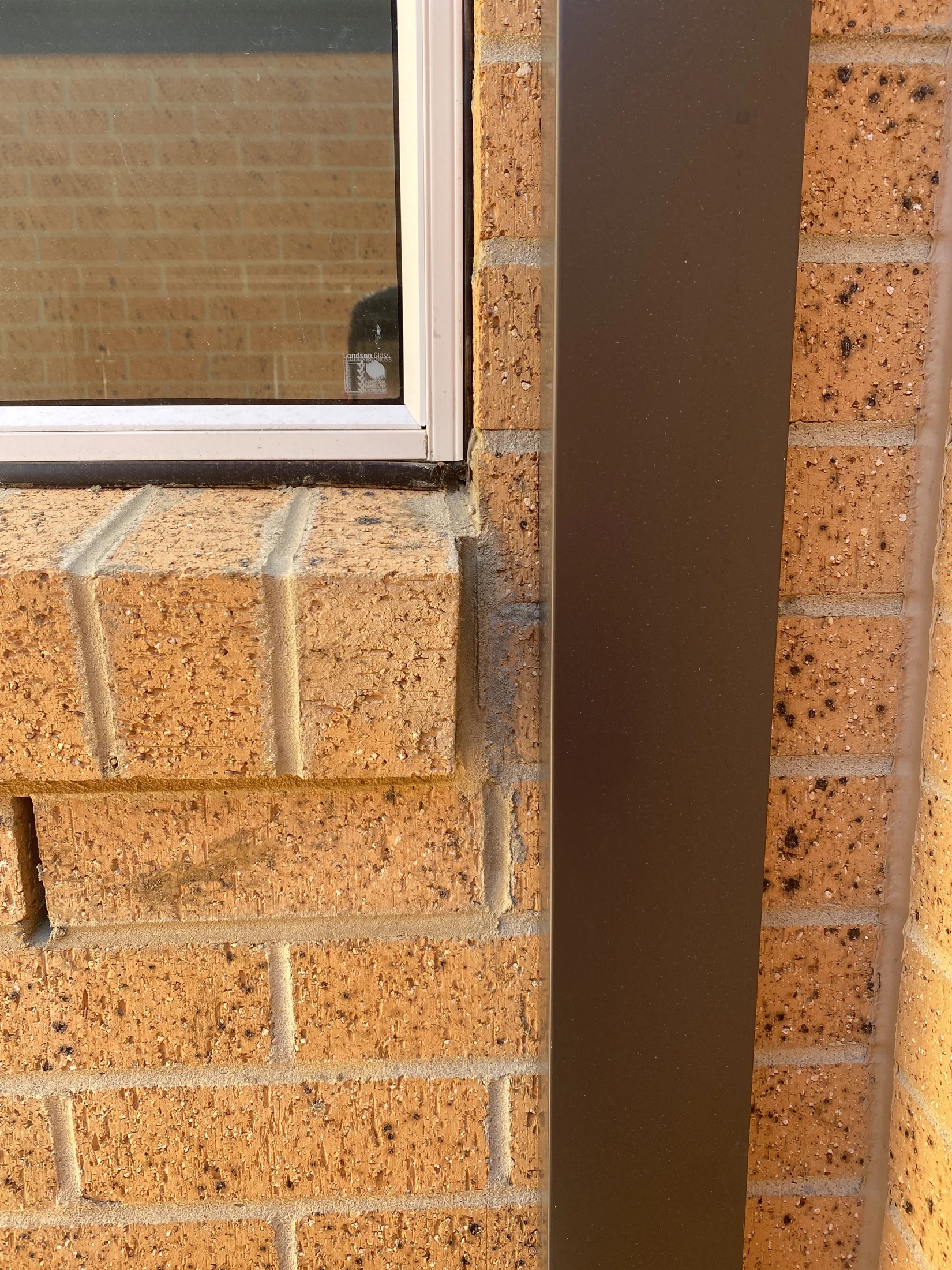 Close-up of a brick wall with a metal edge and window. The bricks are orange-brown. A small sticker on the window is partially visible.