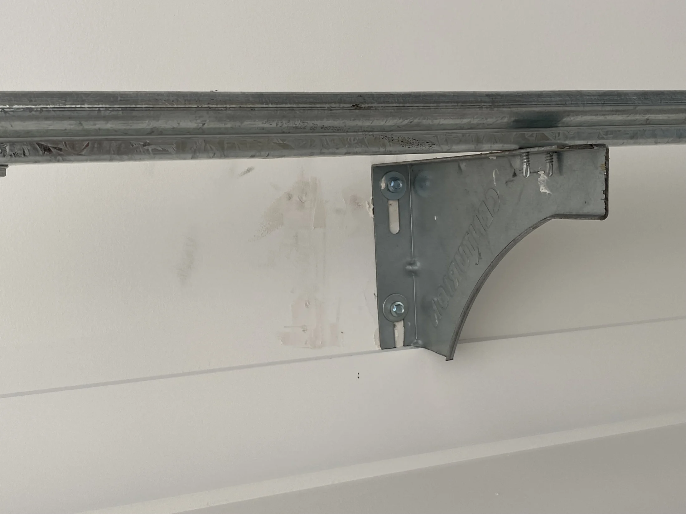 Metal bracket holding a rail against a white wall. The bracket has visible screws and an engraved texture. The setting is a plain interior.